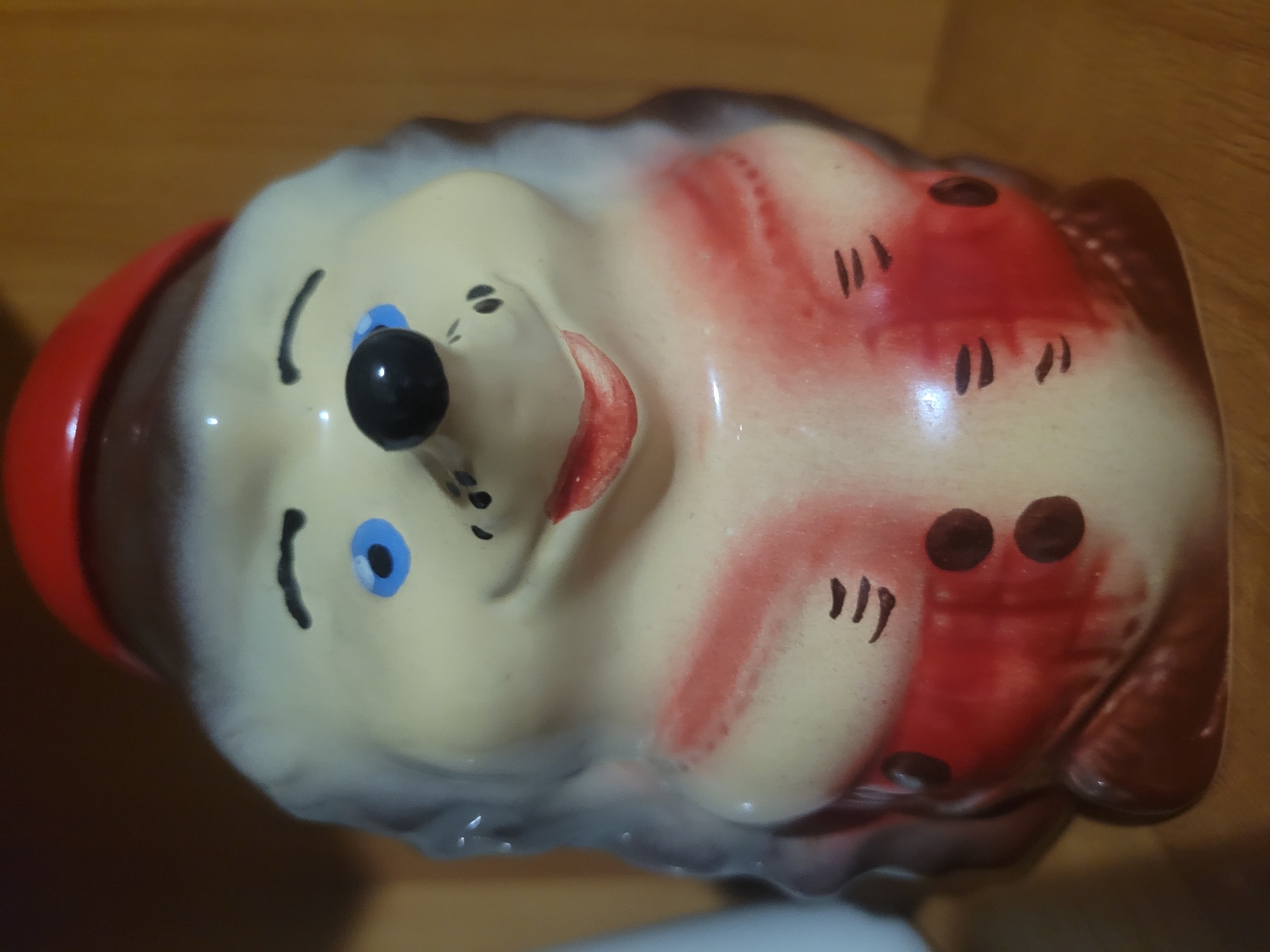 This is a medium shot photograph of a ceramic figurine of a hedgehog. The figurine is beige with gray spines, a red hat, blue eyes, a black nose, and red lips. It is wearing a red jacket with two dark brown buttons on each side. The figurine is positioned in the center of the frame, against a blurred wooden background. There is no discernible location in Belaazyorsk, Belarus.