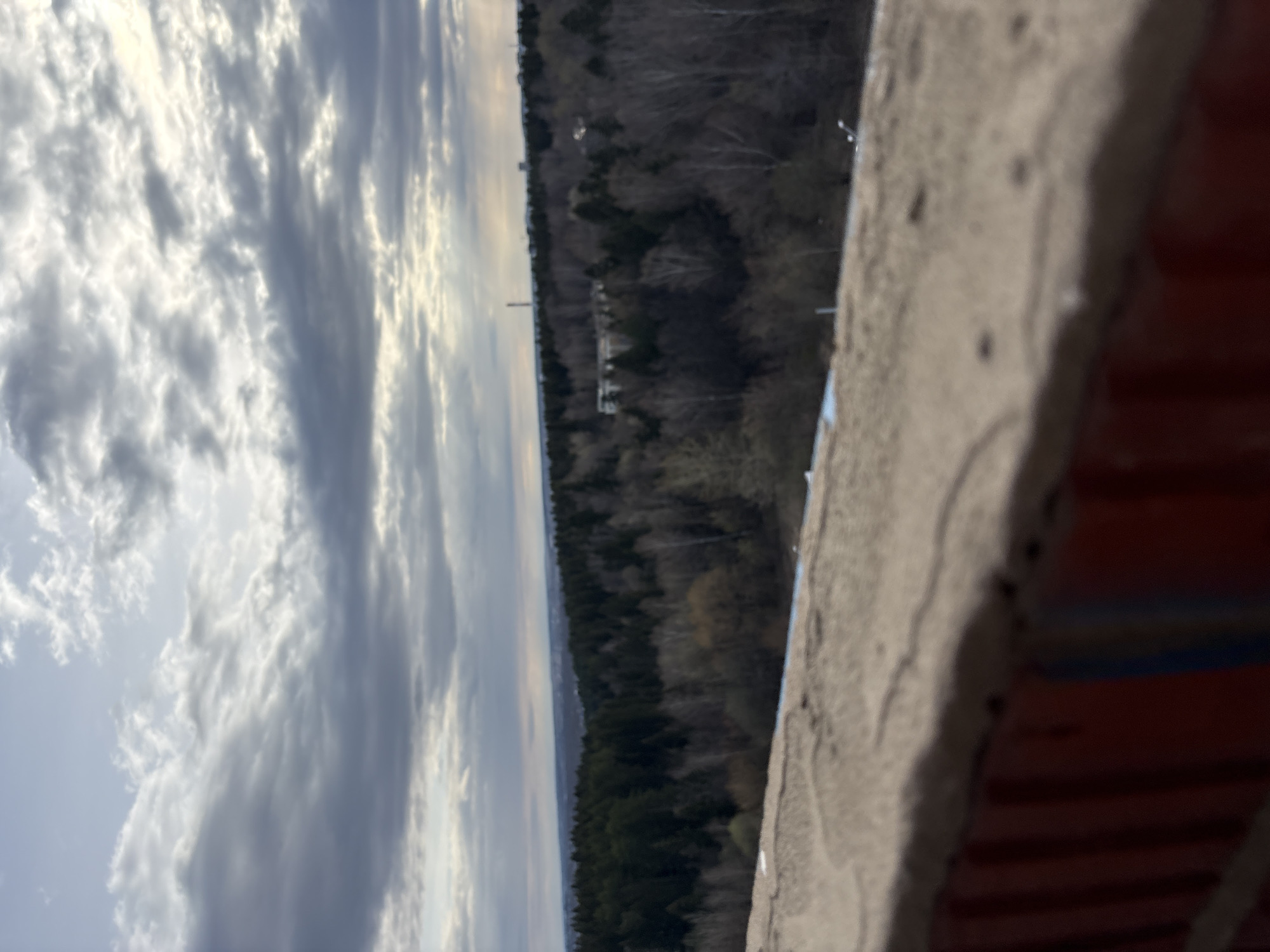From a high vantage point, possibly a tower, a view unfolds encompassing the sky, a forest, and architectural elements. The sky is filled with clouds, casting shadows and highlights. The forest, dense with trees, stretches towards the horizon. In the foreground, a wall with brick and concrete is visible. Given the information that this image is within the city of Izhevsk, Russia, the scene suggests a view from one of the higher structures in the city. There is no human activity explicitly depicted in the image.