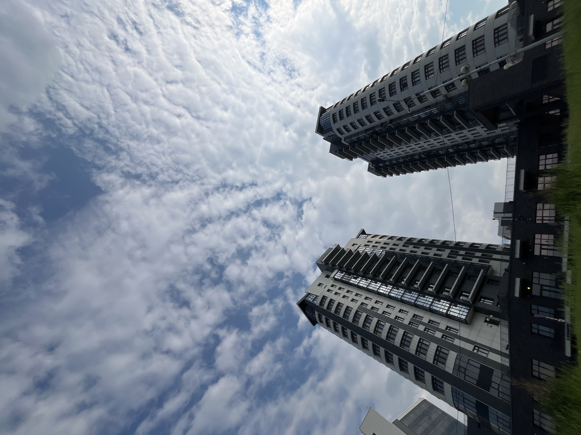 The image shows two modern high-rise apartment buildings in the city of Krasnoyarsk, Russia. The buildings are tall, with curved facades and numerous balconies. The architecture is contemporary, featuring a mix of light-colored panels and dark-tinted windows. The sky above is partly cloudy, with white, wispy clouds scattered across a blue background. The perspective is from below, looking up at the buildings, emphasizing their height. The lower portion of the image shows the dark-colored base of the buildings, with rows of windows. In the foreground, there is a strip of green grass. No people are visible in the image. The scene appears to be during daylight, under a bright sky.