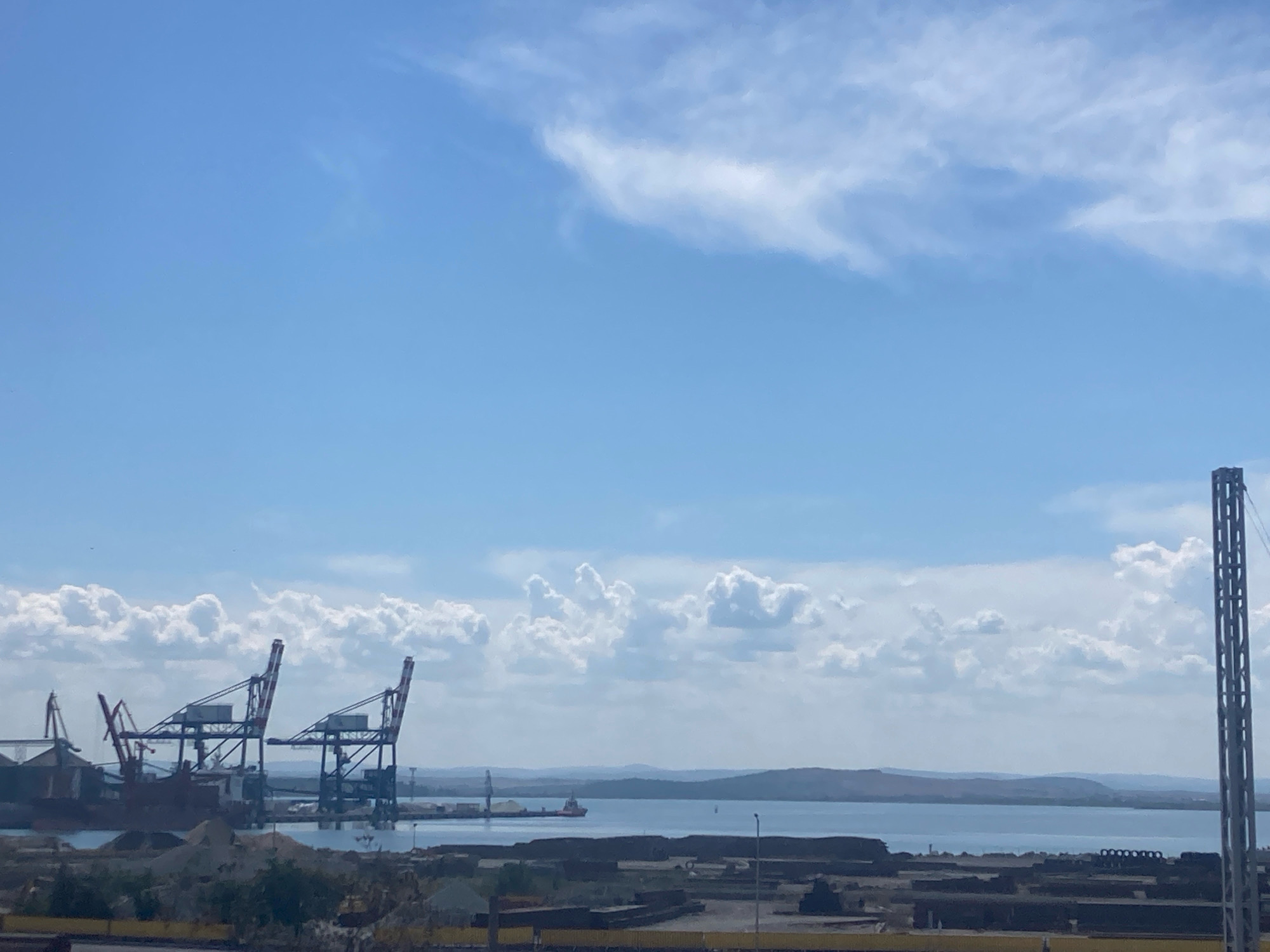 This is a photograph of a port facility on a bright, sunny day, likely taken in Burgas, Bulgaria. The sky is a clear blue with scattered white, fluffy clouds, some of which are wispy and stretched across the upper portion of the frame. In the middle ground, a calm body of water, presumed to be the sea or a large bay, stretches towards a distant, hazy shoreline with rolling hills.
Dominating the left side of the mid-ground are several large industrial cranes, characteristic of a port. Two prominent, tall cranes with red and white detailing are positioned side-by-side, their booms extended. To their left, another, red crane is visible, along with a large industrial building or silo.
In the foreground, the land appears to be a mix of industrial development and construction materials. There are piles of dirt or sand, as well as what seem to be dark-colored, flat structures or shipping containers. A very tall, slender, metal tower with a grid-like structure stands on the far right edge of the image, rising from the foreground industrial area.
A small tugboat or working vessel is visible on the water, to the right of the central cranes. The overall scene depicts an active industrial port with elements of heavy machinery, maritime activity, and a backdrop of natural landscape under a bright sky. There are no people visible in the image.