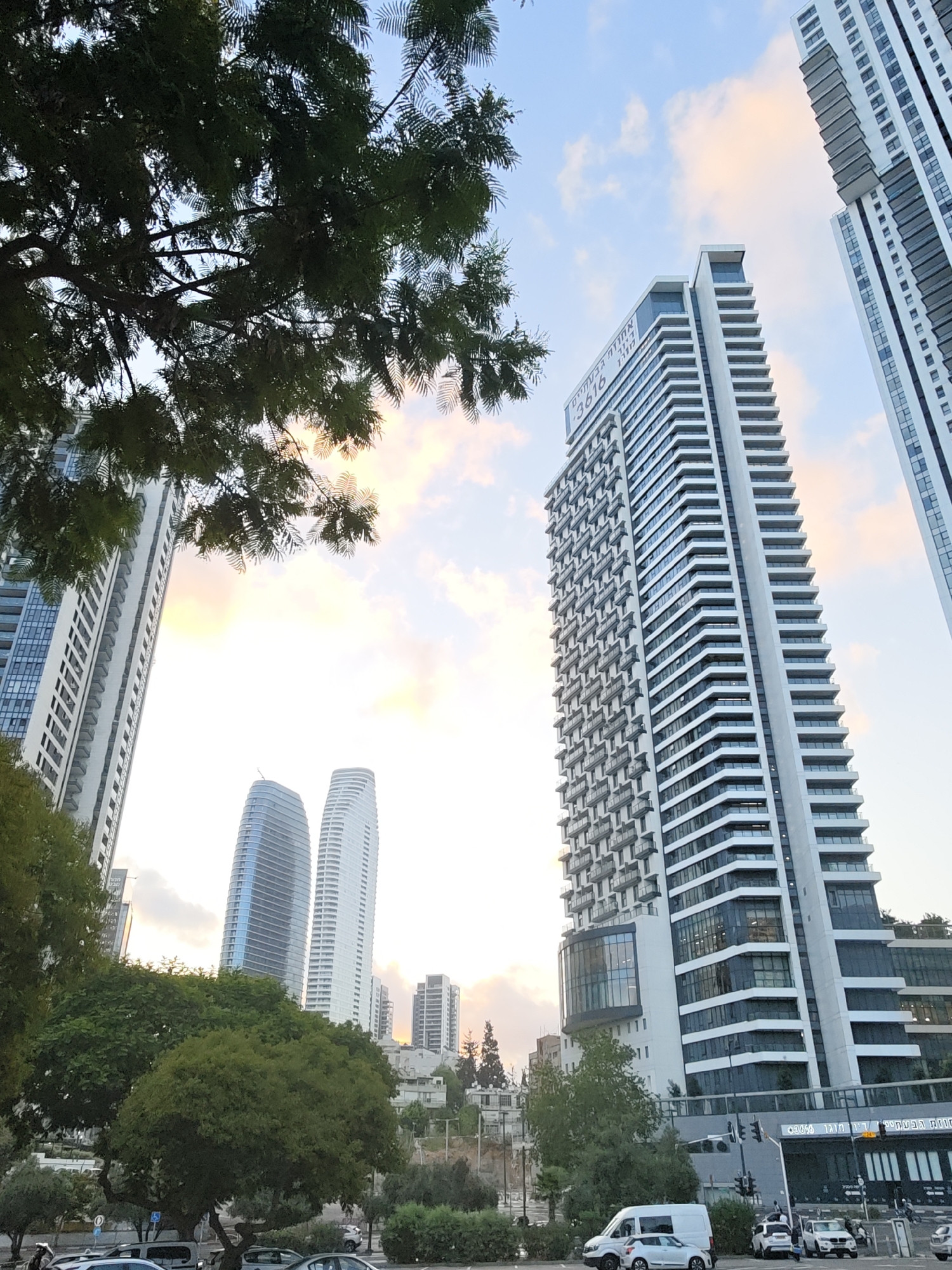 The image depicts a street scene in Tel Aviv, Israel, featuring modern high-rise buildings and lush trees. A prominent tall, white skyscraper with a distinct staggered facade and numerous balconies dominates the center of the frame, with an advertisement featuring the numbers "3616" and Hebrew text visible on its upper section. To its left, another white skyscraper with a more traditional, straight design is partially visible. In the background, two curved, glassy skyscrapers with a blue tint stand out. The foreground is occupied by green trees, some with dense foliage, and a street with several vehicles, including a white van and two cars, parked or driving. Traffic lights and street signs are also present. The sky is partly cloudy with soft, warm hues, suggesting it might be late afternoon or early morning. A few pedestrians are visible on the sidewalk near the street. The scene is a blend of urban architecture and natural elements, characteristic of a modern city.