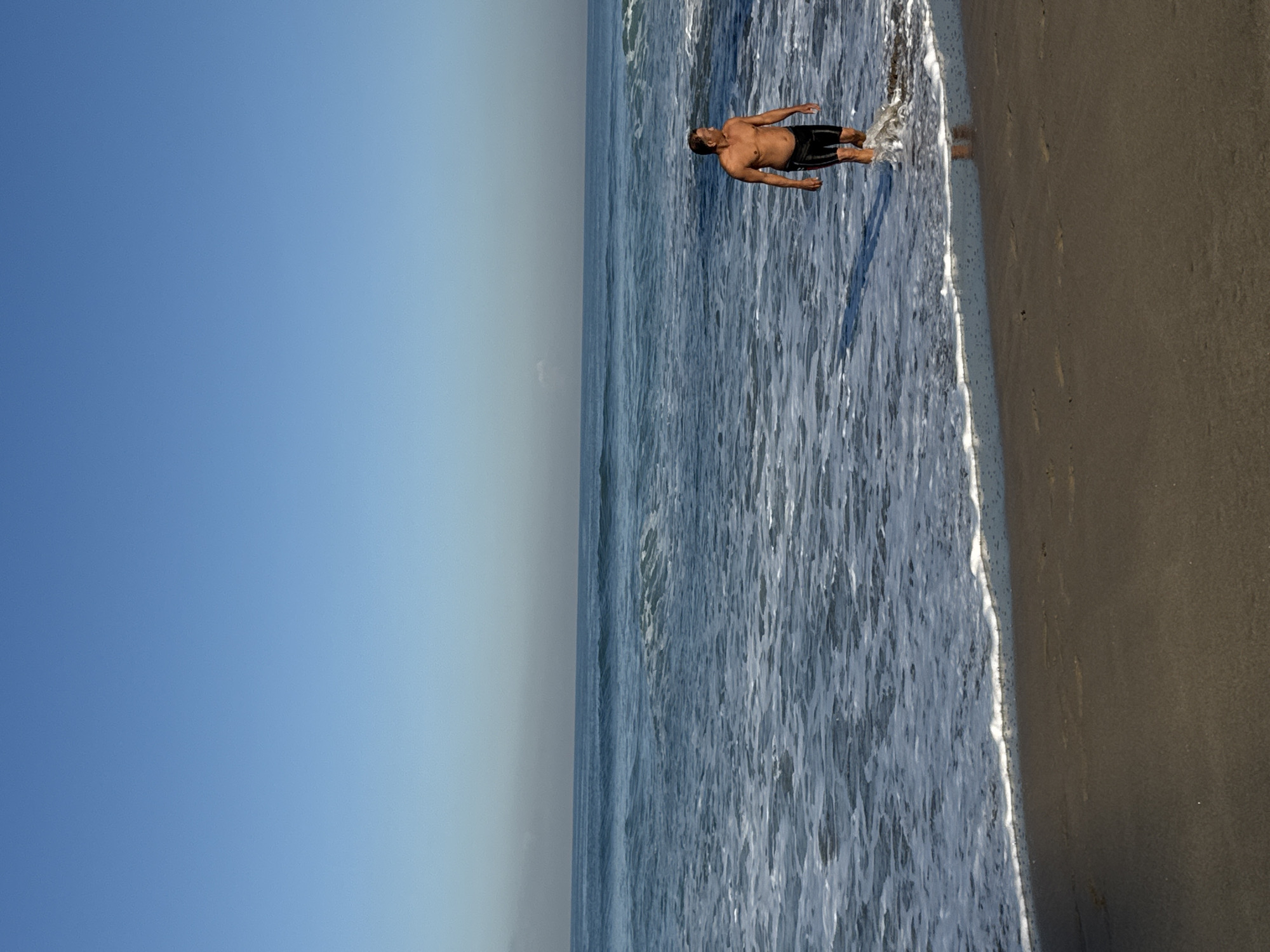 The image depicts a man standing in the surf along a beach. The sky is clear and blue. The water appears to be slightly turbulent with visible foam and waves near the shoreline. The beach sand is dark. The man is standing in the water near the beach.
There are no elements in the picture to confirm this is Kuta, Indonesia.