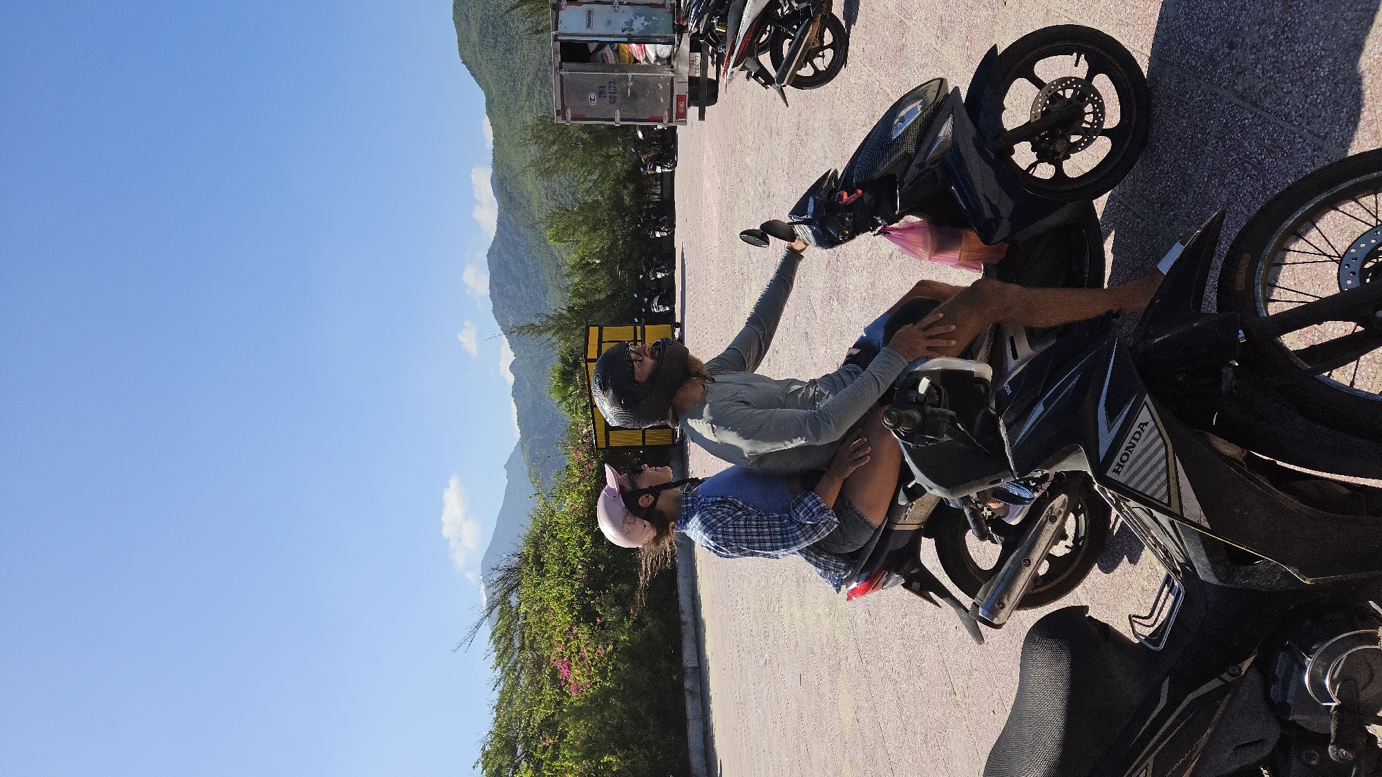 A couple wearing helmets sits on a parked Honda motorcycle in a parking area in Cam Lam, Vietnam. The woman wears a pink helmet and a blue plaid shirt. The man wears a black helmet and gray shirt and is holding the motorcycle's mirror. Behind them is a truck, more parked motorcycles, trees, a clear blue sky, and mountains. The parking area is paved with red and gray tiles.