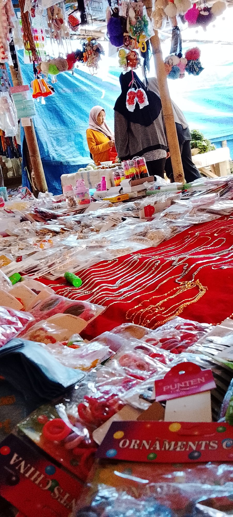 An outdoor market stall is depicted, likely in Bahway, Indonesia, with a variety of goods laid out for sale. The foreground is dominated by numerous small, individually packaged items, many appearing to be jewelry or accessories like necklaces and rings, presented in clear plastic packaging. A red fabric with intricate silver and gold embroidery is visible, partially covered by the packaged goods. In the midground, a table displays more items, including small plastic containers with colorful contents, and bottles of pink liquid. Behind this, two individuals are partially visible. One, a woman wearing a light pink hijab and an orange long-sleeved shirt, appears to be attending to the stall. The other person, dressed in a gray hoodie and black hijab, stands slightly further back. The stall is set up under a blue tarp, supported by wooden poles, suggesting a temporary or semi-permanent market setup. Various hanging decorations, some resembling pom-poms and keychains, are suspended from the stall's structure. The lighting suggests it is daytime.