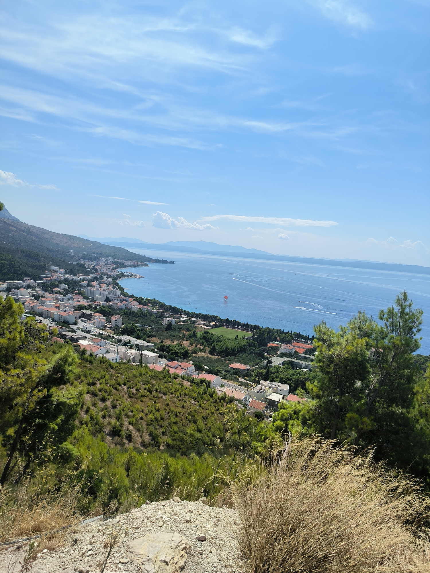This image depicts a high-angle, panoramic view of a coastal town nestled along a mountainside overlooking the Adriatic Sea. The scene is bathed in bright sunlight under a clear blue sky with scattered wispy white clouds.

In the foreground, the terrain is rough and rocky, covered with dry grasses and sparse green bushes, suggesting a natural hillside. Two prominent evergreen trees with dense green foliage are visible on the right and left sides, framing the view.

The middle ground reveals the town, characterized by densely packed buildings with white walls and red-tiled roofs. The architecture appears to be a mix of residential and some larger, more modern structures, possibly hotels or apartments. A football field or sports complex with green turf is noticeable amidst the urban sprawl.

The background is dominated by the expansive, deep blue Adriatic Sea, stretching towards the horizon. Several boats are visible on the water, leaving faint wakes, and a small red parachute is being towed behind a boat, indicating recreational activity. The distant coastline is lined with mountains, softened by atmospheric haze, under the vast sky. The location is identified as Općina Tučepi, Croatia.