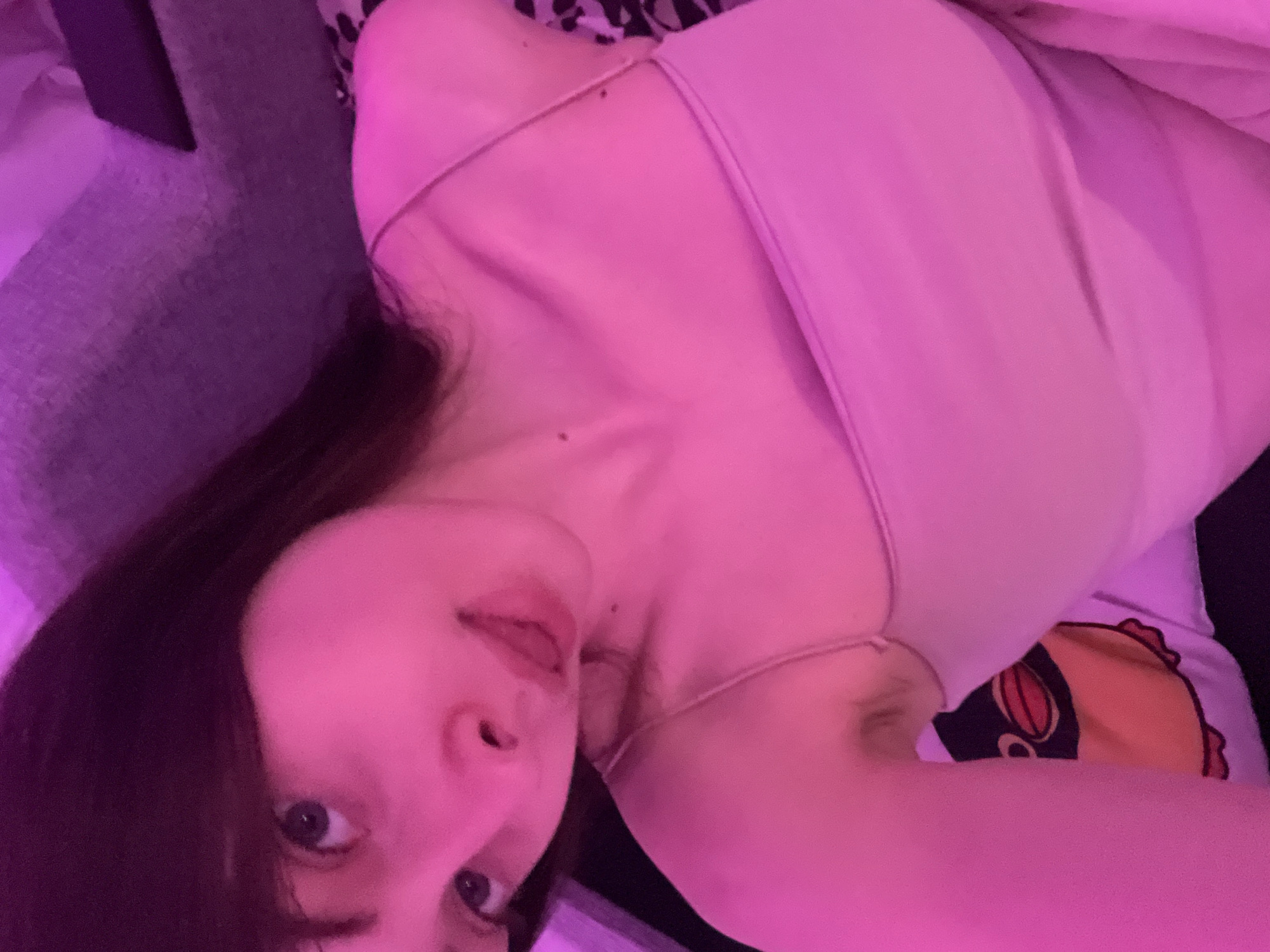 A woman lies on a patterned bed with a pillow that has a cartoon character on it. The light in the room has a pink or purple hue. The woman has dark hair and blue eyes and is wearing a sleeveless top. The location is not identifiable as Saint Petersburg, Russia.