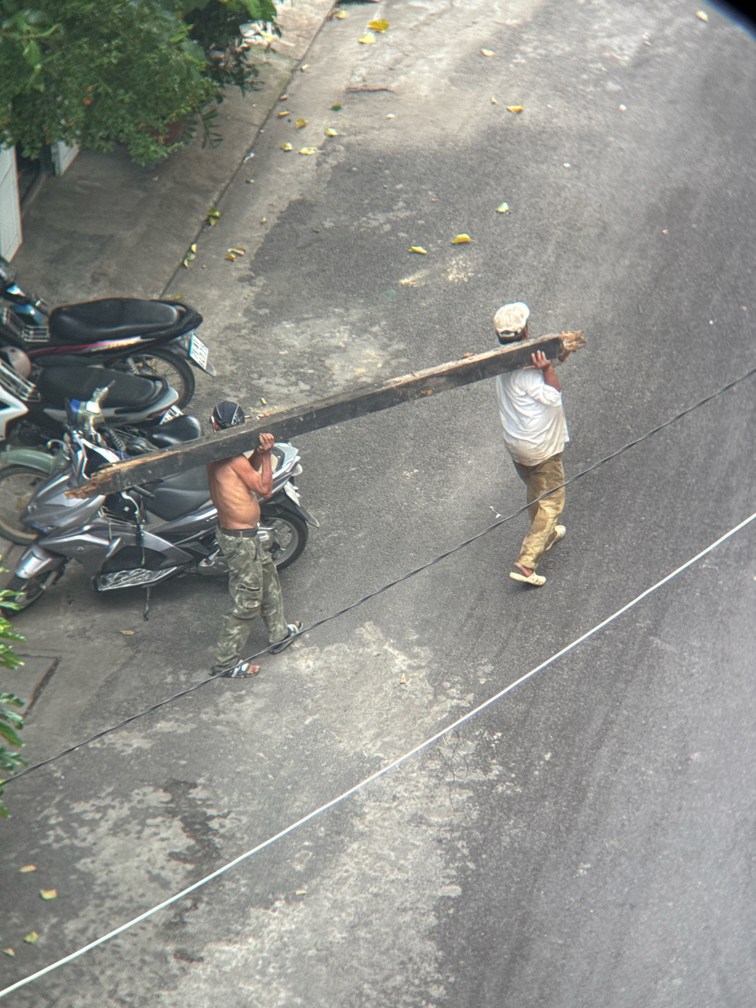 The image, captured from a high angle, shows two men carrying a large wooden beam on a street in Nha Trang, Vietnam. The street is paved with asphalt and shows signs of wear and tear, with scattered leaves and debris. To the left of the street, a sidewalk is visible, adjacent to a building and lush green foliage. Several motorcycles are parked along the sidewalk.
The man in the foreground, on the left, is shirtless and wearing camouflage pants and sandals. He is carrying the beam on his right shoulder. The man behind him, on the right, is wearing a white t-shirt, light brown pants, and a white baseball cap, also carrying the beam on his shoulder. Both men appear to be working together to transport the heavy object. The overall scene depicts a mundane, everyday activity taking place in an urban environment.