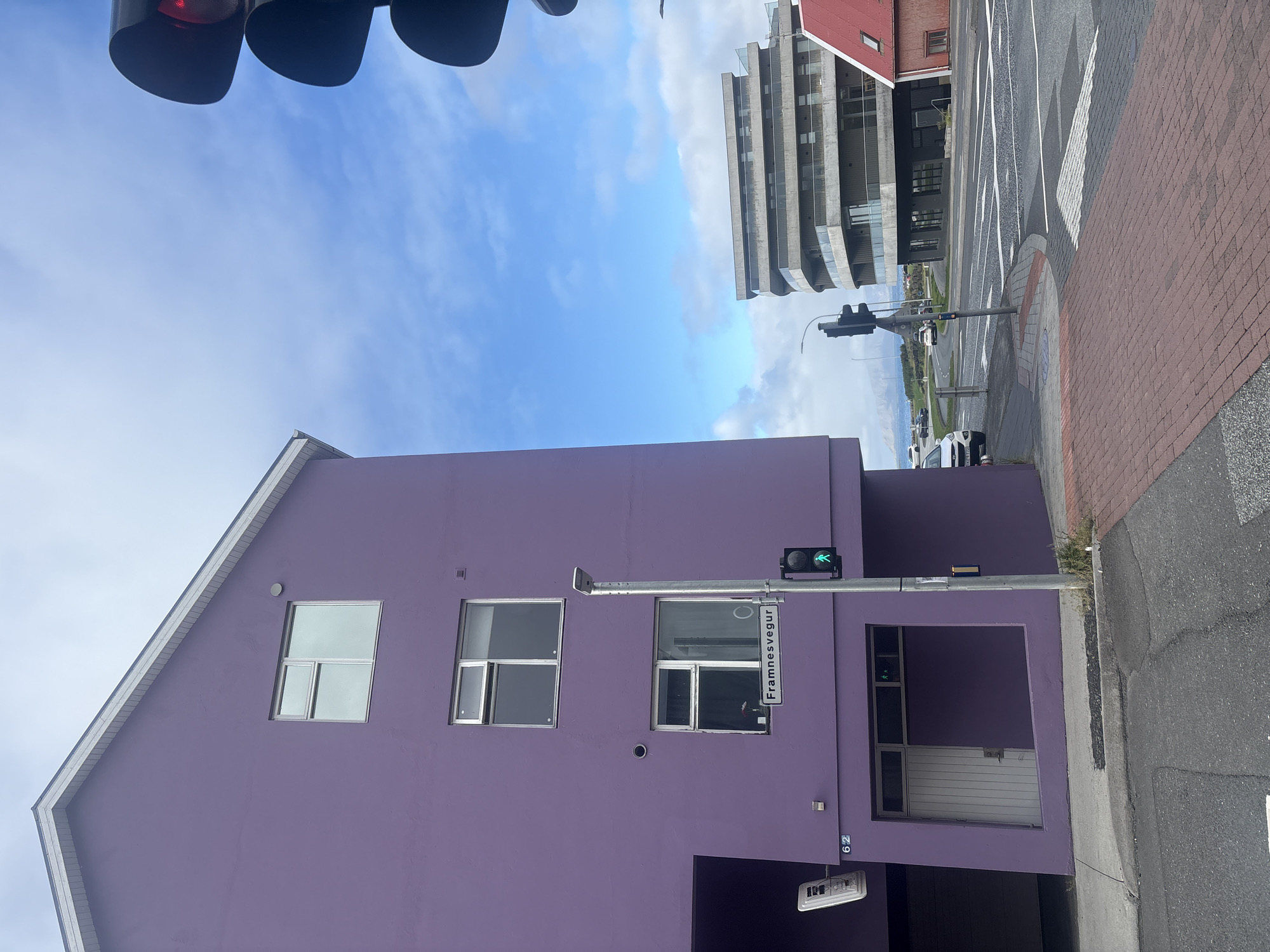 A vibrant purple building dominates the left side of the image. It has a pitched roof and several windows. Below the windows, a white door is visible, and to the left of the door, a sign reads "62". A street sign with the text "Framnesvegur" is mounted on a pole next to a traffic light showing a green pedestrian signal.
To the right of the purple building, a modern, multi-story building with a curved facade and glass balconies stands prominently. Adjacent to it, a red-roofed building with small windows is visible. The scene appears to be an urban intersection in Reykjavik, Iceland, with roads, sidewalks, and other vehicles present. The sky is partially cloudy.