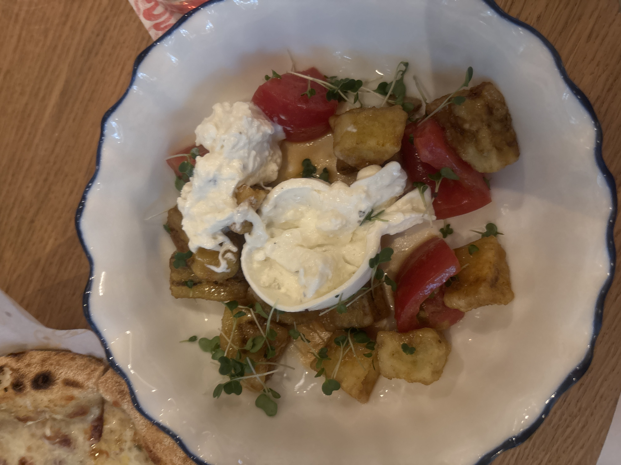 In Novaya Usman', Russia, a white scalloped bowl with a blue rim contains a dish of fried potato cubes, chopped tomatoes, microgreens, and a dollop of creamy white cheese, likely ricotta or burrata. The bowl sits on a wooden table. To the left, a portion of a pizza with melted cheese and visible crust is visible. The lighting is natural and bright, suggesting an indoor setting during the day. No people are visible in the image.