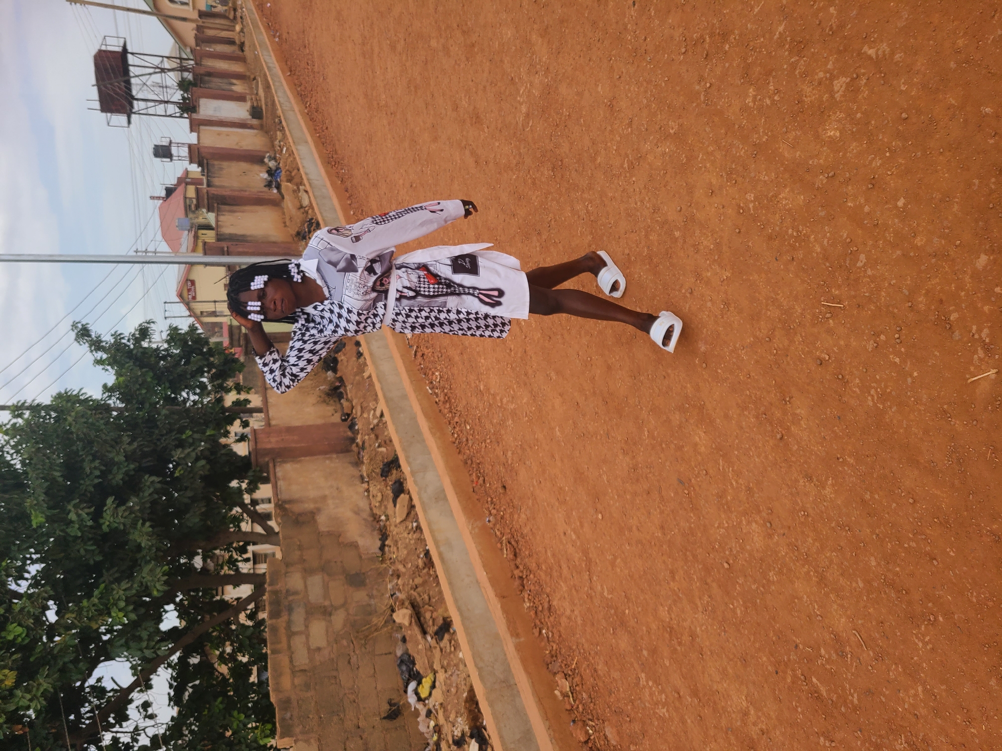 A young woman stands on a red dirt path in what appears to be a neighborhood setting. She is wearing a black and white patterned outfit with white slides. Her hair is styled in braids adorned with white beads. Her right hand touches her head, seemingly posing for a photograph. Behind her, there's a line of buildings, trees, and utility poles. Debris is visible near the curb. The general location is likely the city of Jos, Nigeria based on the architecture and landscape visible.