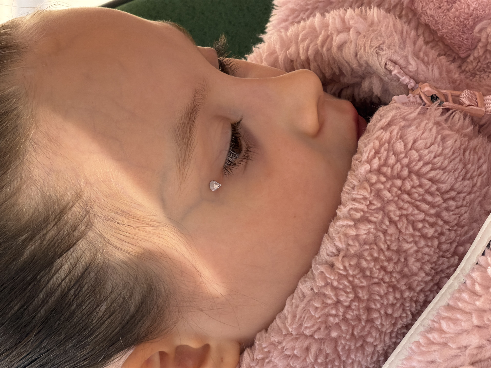A close-up of a young child lying down, possibly asleep. The child's face is in profile, with their nose and mouth visible. A small, teardrop-shaped jewel is visible below their eye. The child is partially covered by a pink, fluffy jacket with a visible zipper. A small portion of green fabric is visible in the upper left corner of the image. The location of the image is unknown and cannot be identified as Perm, Russia.
