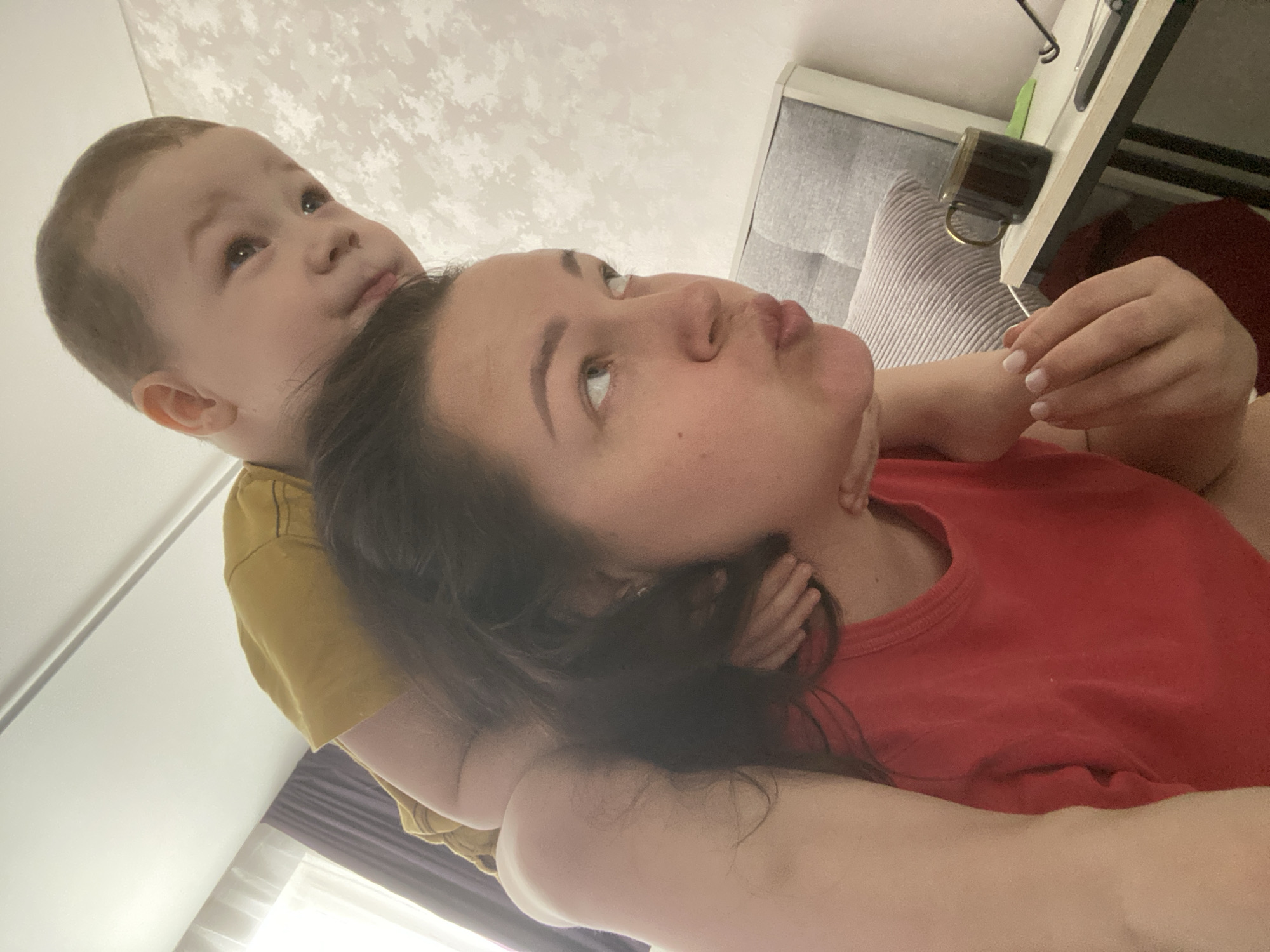 A woman wearing a red shirt is lying down, and a young child wearing a yellow shirt is positioned behind her, looking upwards. The woman is also looking upwards. The background includes a window, a couch, and a table with a mug. The location is unspecified, and there is no evidence to suggest it is in Sochi, Russia.