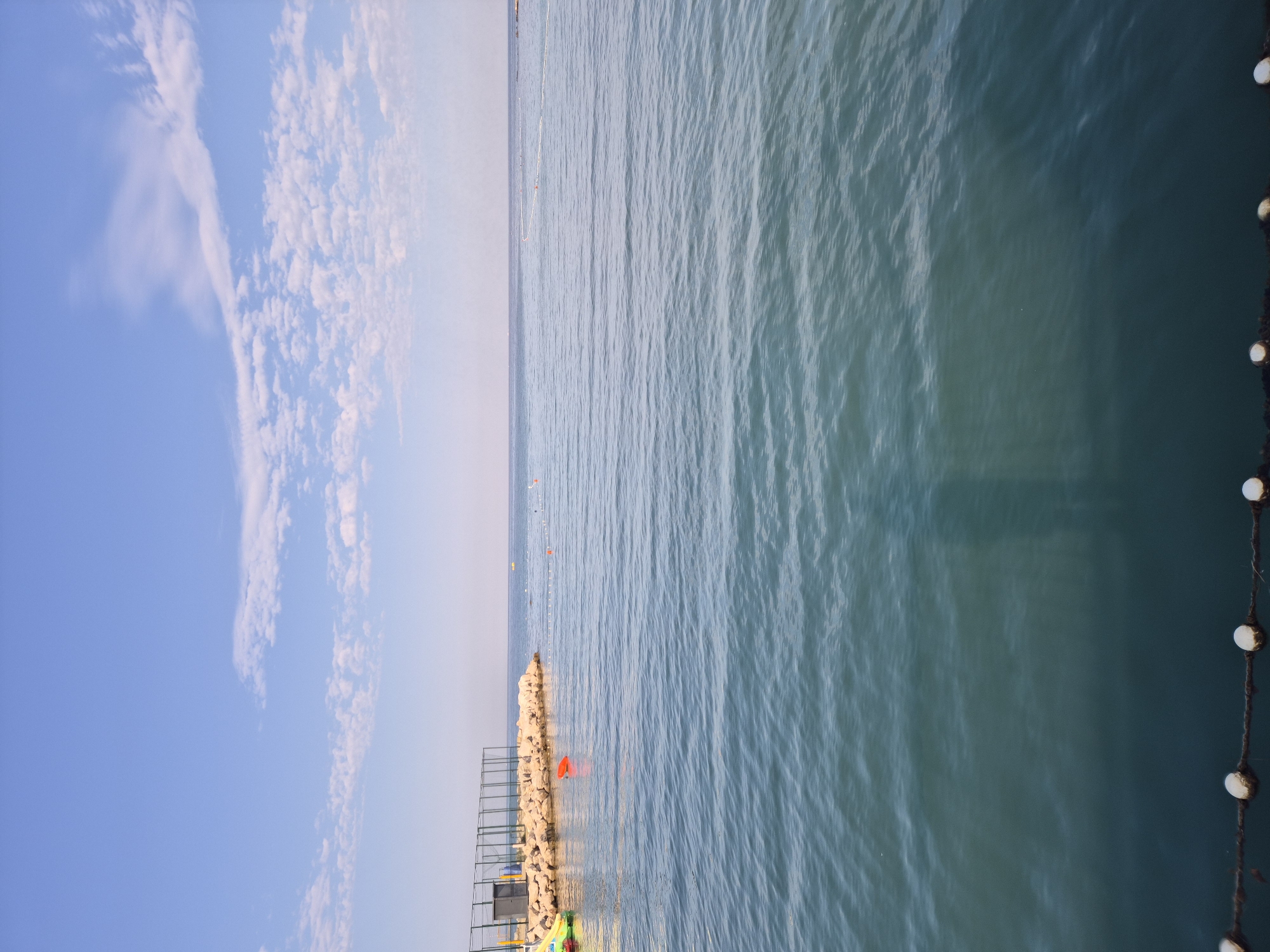 The image depicts a serene seascape with calm turquoise water under a bright blue sky with scattered white clouds. In the distance, a rocky pier with a fence and a small structure is visible, along with a few buoys in the water. The foreground shows the water extending to the bottom edge of the frame, with a rope line featuring white floats marking the edge of a swimming area. There are no people visible in the image. The scene appears to be a tranquil beach or waterfront area, possibly in Umag, Croatia, a coastal town known for its beautiful Adriatic Sea views and recreational areas.