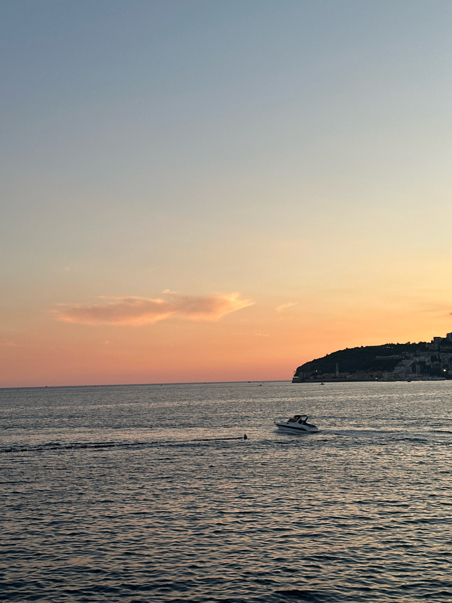This image depicts a serene sunset over the Adriatic Sea in Budva, Montenegro. A white motorboat with a dark trim is navigating the choppy water, leaving a visible wake. In the background, the silhouette of the Budva coastline is visible, characterized by a prominent hill covered in lush vegetation and scattered buildings that appear illuminated. The sky exhibits a gradient of colors, transitioning from a pale blue at the top to soft hues of orange and pink near the horizon, with a few wispy clouds present. The sea itself is a deep blue, reflecting the colors of the sky and appearing slightly textured due to the gentle waves. A buoy is visible in the water to the left of the boat. The scene conveys a peaceful, late-day atmosphere.