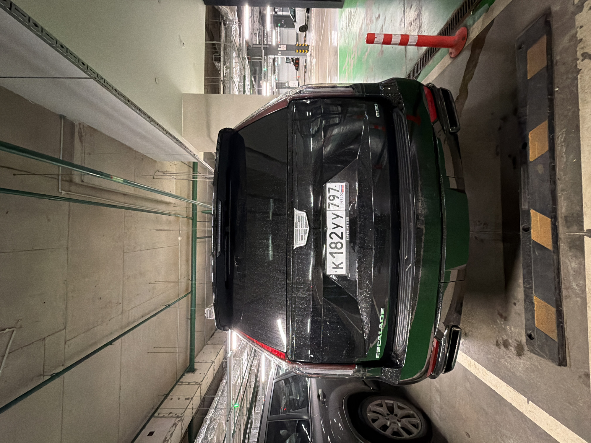 The image depicts the rear of a black Cadillac Escalade parked in an indoor parking garage. The vehicle has a Russian license plate that reads "K 182 YY 797." The location appears to be Moscow, Russia, based on the "RUS" marking on the license plate. The car is positioned near a parking barrier with black and yellow stripes. Overhead, there are exposed pipes, electrical conduits, and other infrastructure typical of a parking garage. Another vehicle is partially visible near the Cadillac Escalade. The lighting is artificial, suggesting the space is enclosed.