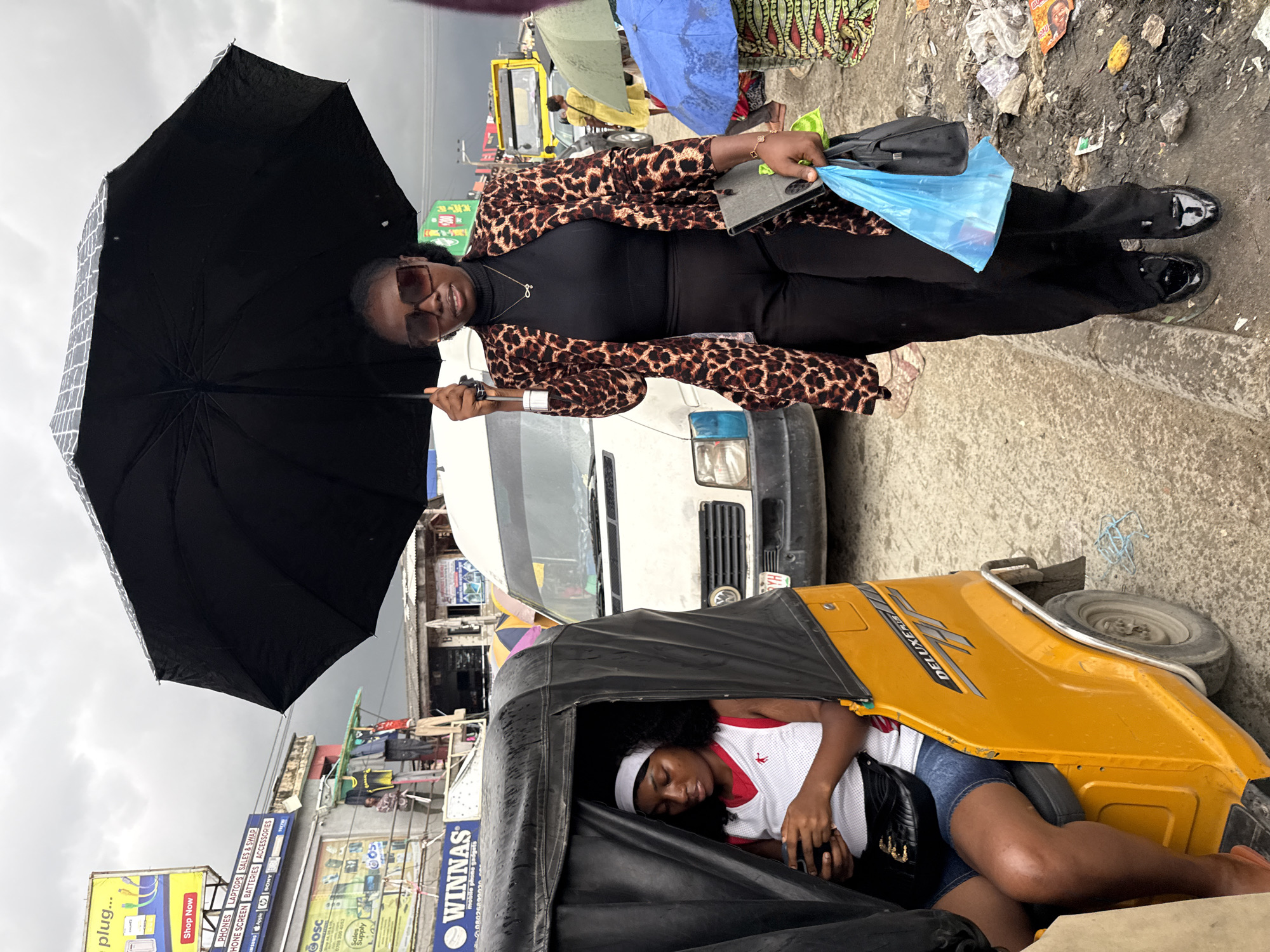 In Lekki, Nigeria, on a cloudy day, a woman stands under a large black umbrella, holding it with her left hand. She wears a leopard-print jacket over a black turtleneck and black pants, with shiny black shoes. In her right hand, she carries a black handbag and a bright blue plastic bag, along with a gray tablet device. She wears large, square sunglasses. To her left, a young woman sits inside a yellow auto-rickshaw, looking down at her phone. She wears a white sleeveless shirt with red trim and denim shorts, and a white headband. The auto-rickshaw has "DELUXE" printed on its side. In the background, a white van is parked, and other vehicles, including a yellow truck, are visible. The ground is damp and littered with debris, including plastic bags and papers. Several other people are visible in the background, some holding umbrellas. The overall scene depicts a busy street with vehicles and pedestrians in an urban environment.