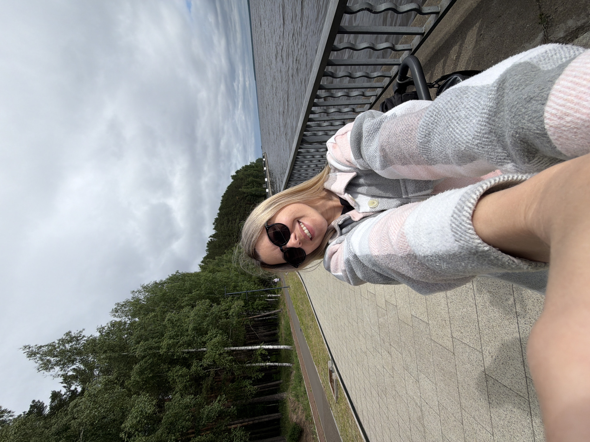 A blonde woman wearing sunglasses and a plaid shirt is taking a selfie in Chaikovsky, Russia. She is standing on a paved walkway next to a body of water. Trees line the walkway to her left, and a metal railing runs along the edge of the water. The sky is cloudy.