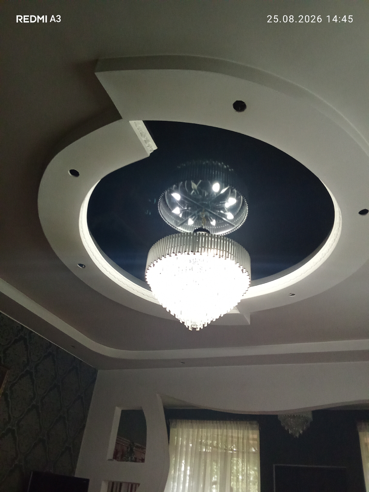 The image shows a view of a ceiling with a large, ornate chandelier. The ceiling has a modern, multi-layered design with recessed lighting and a dark center section. The chandelier is bright and appears to be made of crystal. In the background, there are sheer curtains visible, suggesting an indoor setting, likely a room or hall. The timestamp in the upper right corner indicates the image was taken on August 25, 2026, at 14:45. The text "REDMI A3" is in the upper left corner, indicating the phone model used to take the picture. No people or specific locations within Pshap, Abkhazia, are visible in this image.