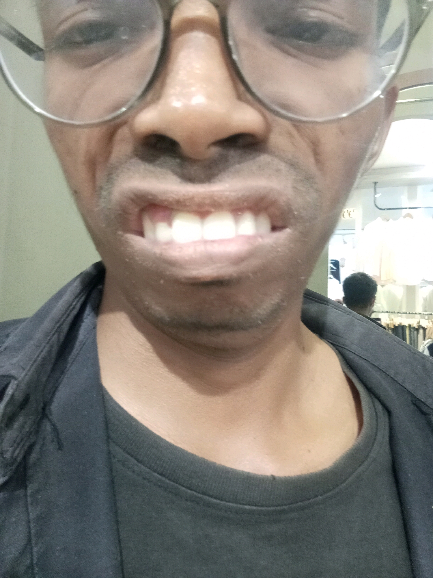 The image shows a close-up, slightly blurry, selfie of a person wearing glasses and a dark shirt, possibly in a clothing store in Surabaya, Indonesia. The person's face is partially visible, with their mouth open showing teeth, and their eyes are visible behind large, round glasses. In the background, there are racks of clothing, suggesting a retail environment. The lighting is bright, and there is a reflection in what appears to be a mirror or glass surface, showing another person's head.
