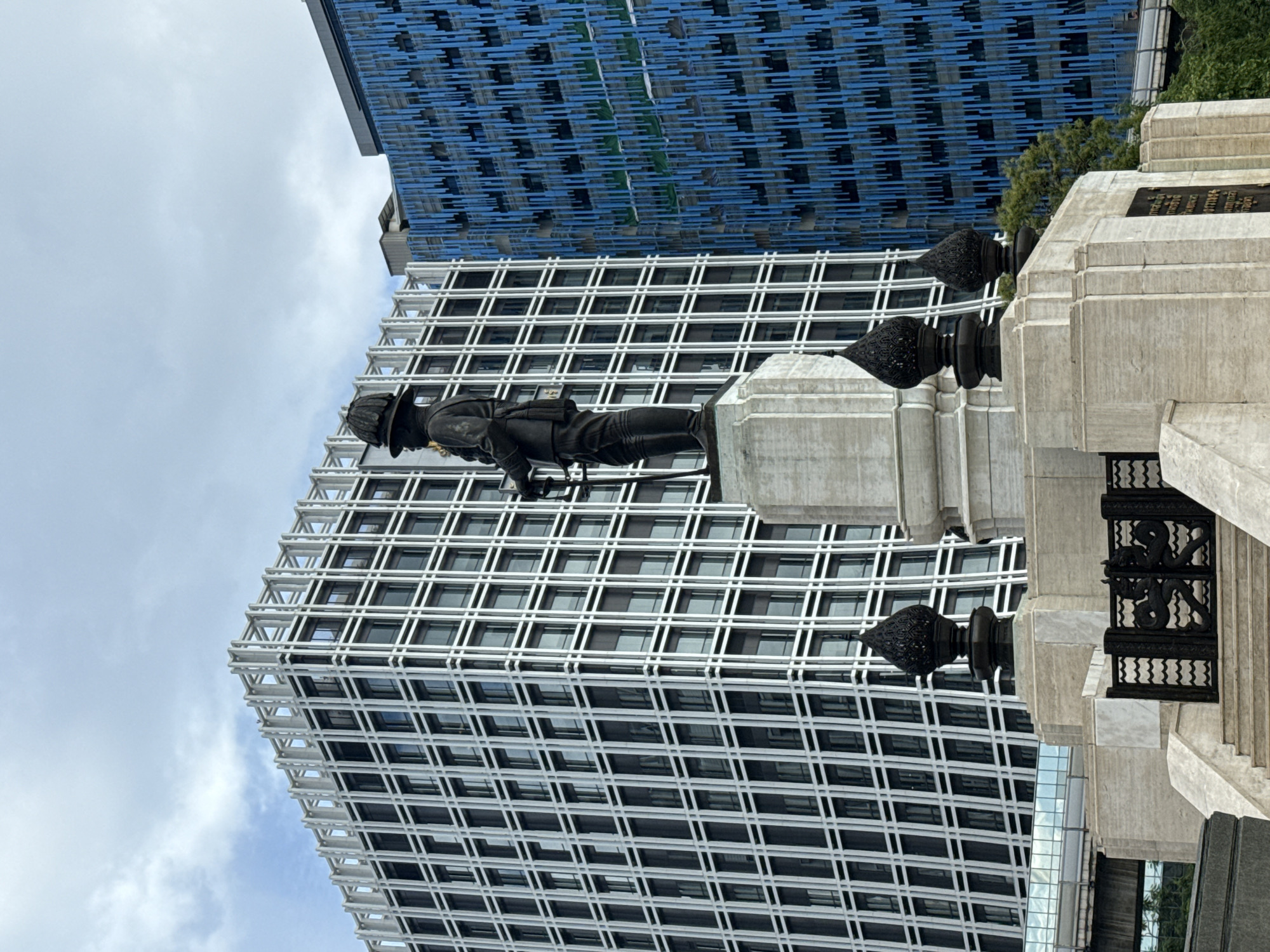 The image features a firefighter statue against a backdrop of modern architecture in Bangkok, Thailand. The statue is a bronze figure of a firefighter in action pose, standing on a light gray concrete pedestal. The pedestal is decorated with dark ornate black metal details and is part of a larger monument with inscriptions. Behind the statue is a towering skyscraper with a white grid-like facade, and to the upper left is a building with a blue linear pattern. The sky is overcast with light gray clouds.