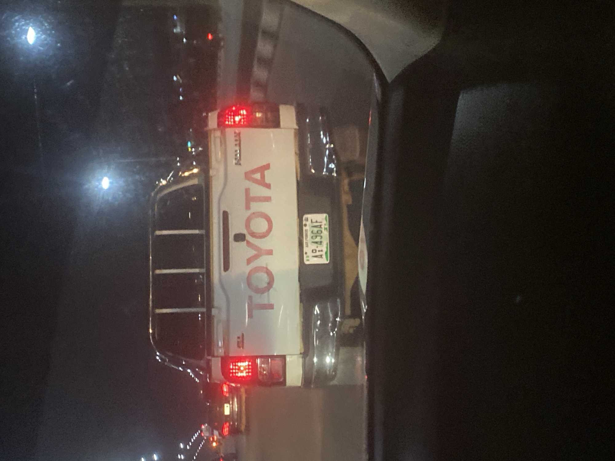 A white Toyota Hilux pickup truck is seen from the rear at night, possibly in traffic, on a paved road with multiple lanes. The truck has a chrome bumper and a visible tailgate with "TOYOTA" written in large red letters. A license plate with the characters "AP-496AF" is mounted below the tailgate. The truck's red taillights are illuminated. In the background, other vehicles with illuminated headlights and taillights are visible, as are streetlights and building lights. The overall scene suggests an urban environment at night, likely on a highway or major road. The provided hints indicate the location is Gosa, Nigeria.