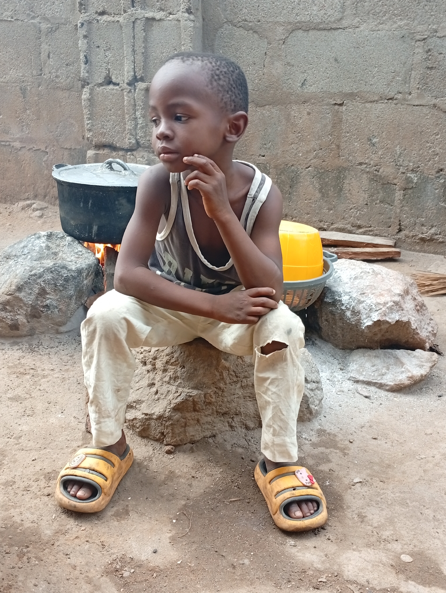 A young Black boy, appearing to be between 4 and 7 years old, is seated on a large rock outdoors in Adankolo, Nigeria. He has short, dark hair and wears a grey and white striped tank top and light-colored trousers with a tear on the left knee. His expression is thoughtful or pensive as he gazes slightly to his right, with his right hand resting on his chin. He is wearing yellow and grey open-toed sandals; a small, white, cartoon-like cat head charm, resembling Hello Kitty, is attached to the strap of his right sandal.
Behind and to the boy's left, a large black cooking pot with a lid is positioned over an open fire, supported by several rocks, indicating outdoor cooking. To his right, a yellow plastic basin is placed within a grey plastic basket. The ground is unpaved dirt or sand, and a concrete block wall forms the background. Further to the right, some wooden planks and a small bundle of sticks, possibly a broom, are visible on the ground. The scene appears to be taking place during the daytime.