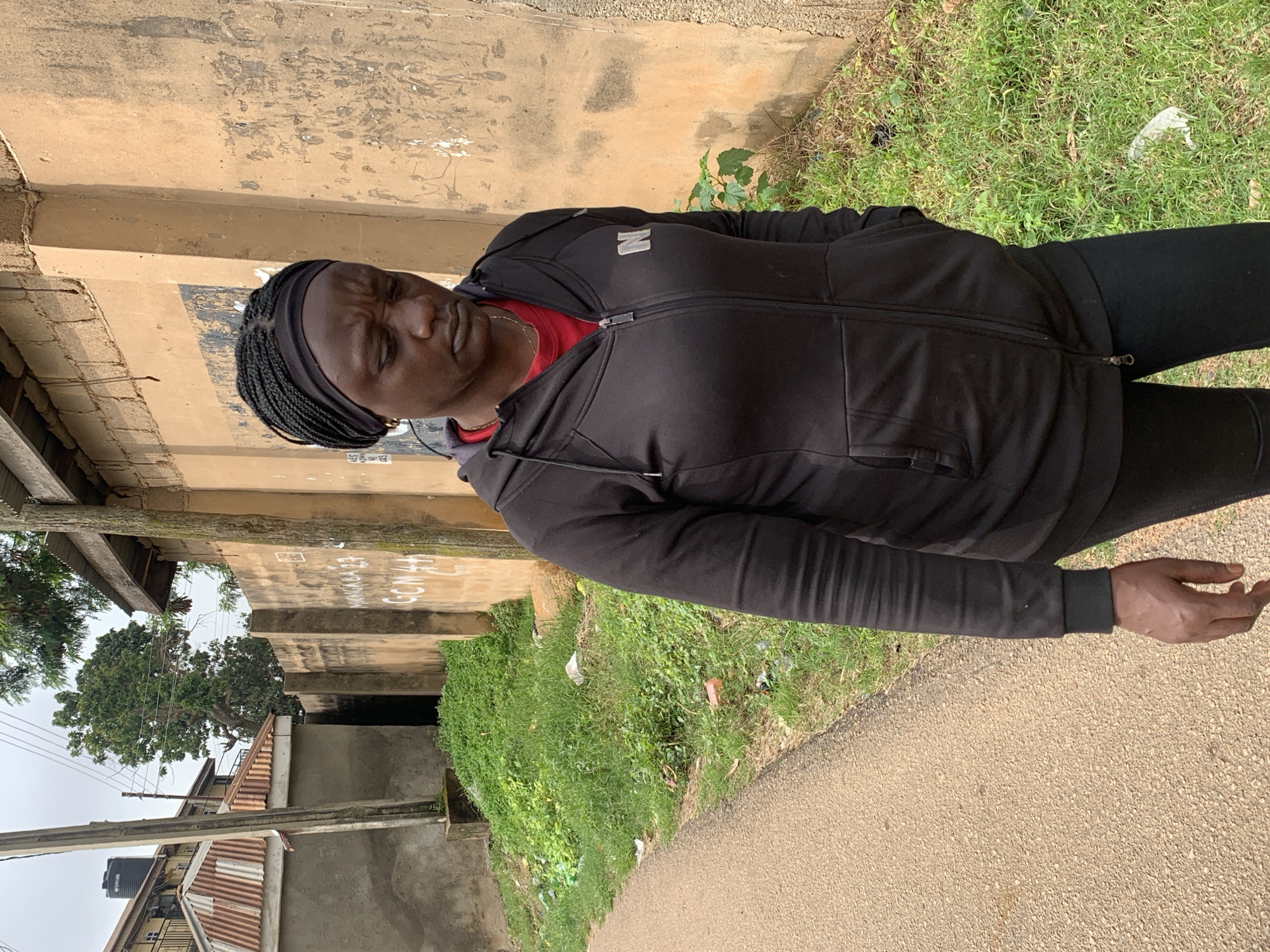 An outdoor medium shot shows a Black woman from the chest up, walking on a dirt path in Jos, Nigeria. The woman is wearing a black hooded jacket over a red shirt, and black leggings. Her hair is braided and she has a black headband on. She is looking down with a concerned expression. The background consists of a textured, light brown wall with some graffiti on it, and a patch of overgrown green grass. Behind the wall, part of a building with a corrugated metal roof can be seen. Overhead, power lines are visible against a cloudy sky.