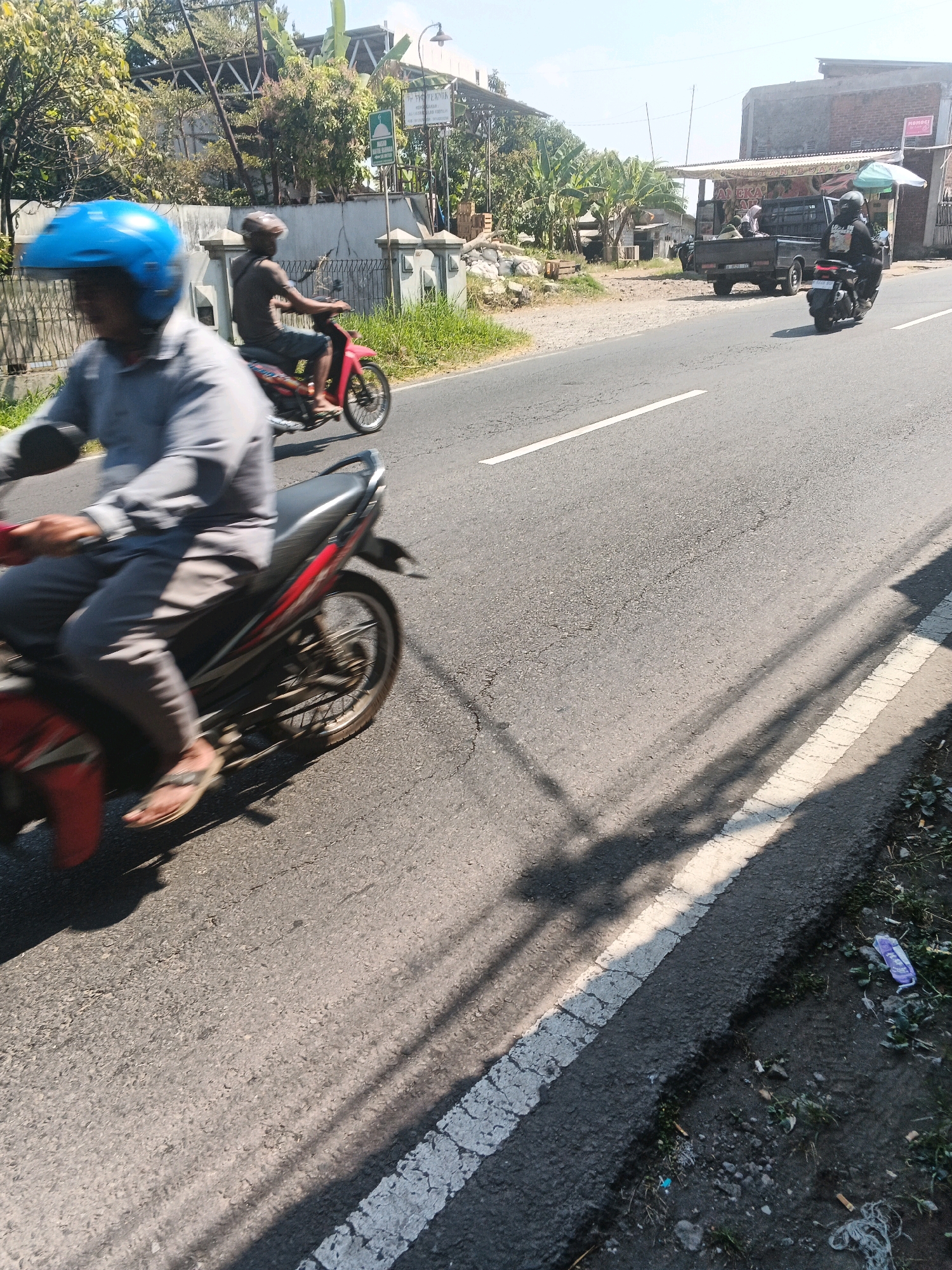 The image depicts a sunlit street scene in Bandungan, Indonesia. In the foreground, a person wearing a blue helmet is riding a motorcycle, their body and the vehicle blurred due to motion. Behind them, another individual on a red motorcycle is also in motion, wearing a dark helmet. Further down the road, a black pickup truck is parked, and a person on a black motorcycle is riding away from it. The street is paved with asphalt and marked with white lines. The environment includes trees, a building with a sign, and a fence. Shadows of trees are cast across the road, suggesting a bright, sunny day. The ground beside the road is covered with sparse vegetation and some litter.