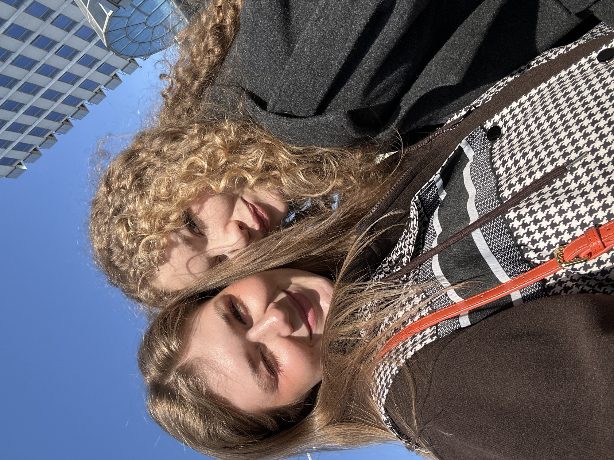 The image depicts two young adult females in a close-up, selfie-style outdoor photograph under a clear blue sky. The female positioned on the left has straight, light brown hair, fair skin, and is seen winking her right eye while smiling. She is dressed in a dark brown zipped garment worn over a black and white houndstooth pattern shirt featuring a horizontal striped design on the upper chest. An orange strap from a bag crosses her upper body. The female on the right has curly blonde hair, fair skin, and a subtle smile, with her face partially obscured by the first individual. She is wearing a dark grey, textured overcoat.
In the upper right background, two contemporary architectural structures are visible. One is a tall, light-colored multi-story building characterized by a repetitive grid of square windows. Adjacent to it, a large, curved glass dome structure is partially discernible. These buildings are consistent with the **Kazan Riviera complex**, specifically the Kazan Riviera Aquapark and Hotel, located in the city of Kazan, Russia.