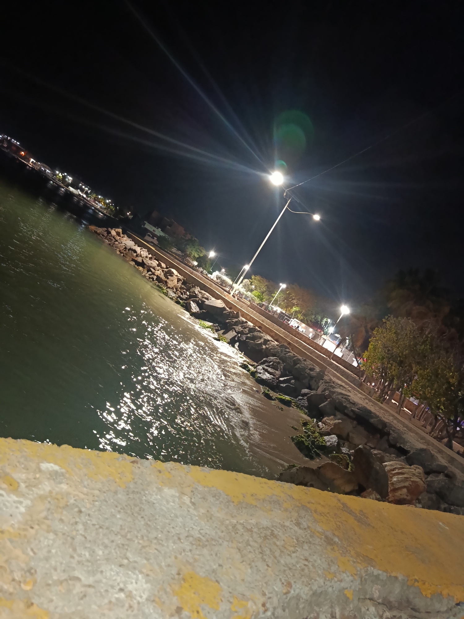 The image depicts a night view of the coastline in Juan Griego, Venezuela. A low wall with peeling yellow paint is in the foreground. Beyond the wall is a rocky shoreline bordering water, possibly the Caribbean Sea. The water reflects light, indicating movement or ripples. Streetlights illuminate a walkway lined with trees and vegetation. In the distance, more lights suggest a populated area along the coast. The angle of the shot is tilted, giving a dynamic perspective of the scene.
