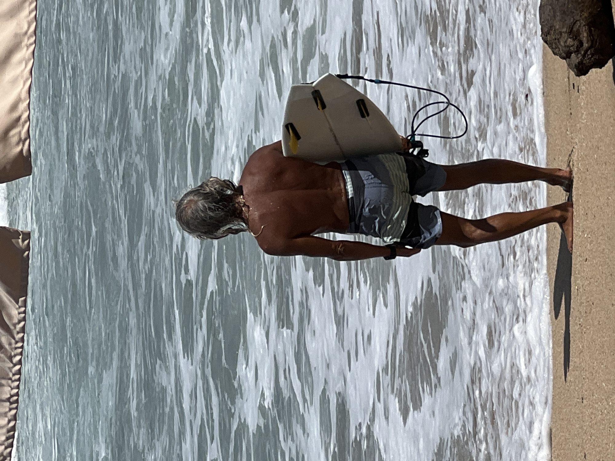 A medium shot captures a man from behind, standing on a sandy beach in Kuta, Indonesia, with his surfboard tucked under his arm. The ocean stretches out before him, its surface textured with gentle waves and white foam lapping at the shore. The man, with his back to the camera, appears to be a surfer, his tanned skin and muscular build suggesting he is ready to enter the water. He wears patterned swim trunks and a dark watch on his left wrist. The surfboard is light-colored with yellow accents, and a black leash trails from it, connecting to his hand. A large, dark rock rests on the sand to the right of the frame. The scene is illuminated by natural daylight, casting shadows on the beach.