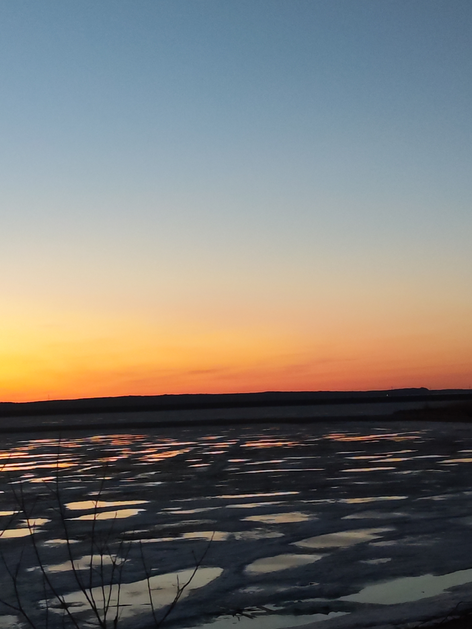 The image depicts a sunset over the Kama River in Chaikovsky, Russia. The sky transitions from blue to orange as the sun sets behind the horizon. The river is partially frozen, with patches of open water reflecting the orange sky. In the foreground, bare tree branches are visible. The land mass in the background is a mix of dark gray and red from the sunset.