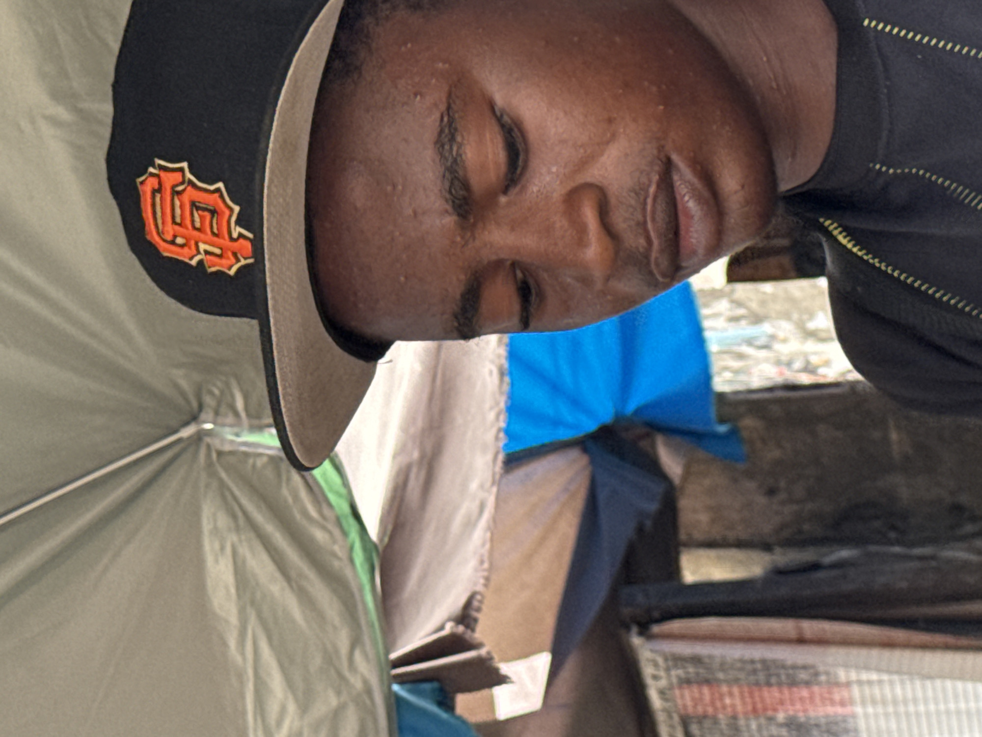 An image, rotated clockwise, depicts a young Black man from a close-up, portrait perspective. He is looking downwards, with his eyes appearing closed or in a relaxed, lowered gaze. He wears a black baseball cap featuring a distinctive orange and beige embroidered "SF" logo, commonly associated with the San Francisco Giants, on its front. The cap has a contrasting light-colored brim. His skin shows some minor texture and small bumps on the forehead. He has short, dark facial hair.
He is dressed in a dark-colored top, possibly black, adorned with subtle, dotted yellowish stripes. The background indicates an informal outdoor or semi-outdoor setting. To the left, a light green or grey fabric structure, resembling a tent or canopy, is visible with internal supports. Further back, on the right, the scene is composed of various makeshift materials, including a bright blue tarp, beige fabrics, and darker elements, suggesting a cluttered urban environment. Portions of a patterned fabric, possibly a rug or mat, are visible at the bottom of the frame. The setting points towards an informal market, street vendor area, or temporary dwelling, consistent with urban landscapes in Lagos, Nigeria. The ambient light suggests it is daytime. The man appears to be in a moment of pause or rest.