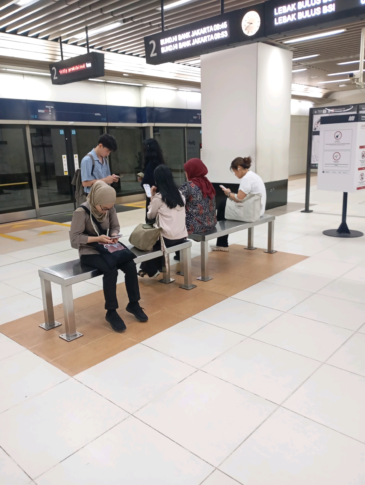 The image depicts a scene at the Bundaran HI MRT station in Central Jakarta, Indonesia. Several people are waiting on benches for a train, with most of them engaged with their mobile phones. In the foreground, a woman wearing a light brown hijab and glasses is seated on a metal bench, looking down at her phone, with a dark tote bag on her lap. To her right, another woman in a red hijab is partially visible, also looking at her phone. Further to the right, a woman in a white t-shirt and headphones is seated, interacting with her phone. In the background, a young man with a backpack is standing and looking at his phone, and two women are also visible, one standing and one seated. The station environment features clean, white tiled flooring, metal benches, and large glass doors on the platform. Overhead, digital signs display train information, including times and destinations like "LEBAK BULUS BSI." The overall impression is one of a typical public transit waiting area, with passengers occupied by their personal devices.
