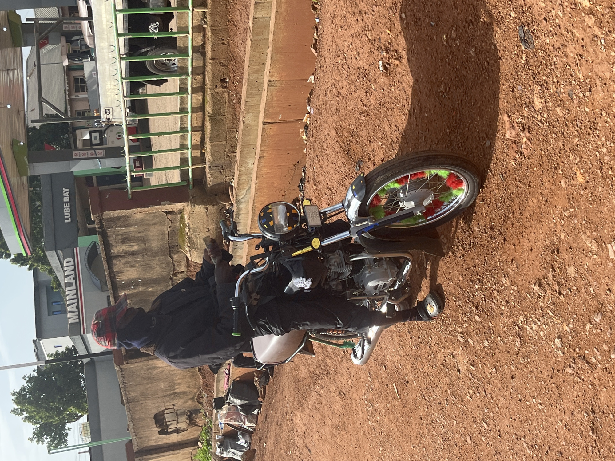 The media file shows a person sitting on a motorcycle. The person is wearing a black jacket, black pants, and a red and blue checkered headwrap. The motorcycle has a decorative wheel with red and green accents. The scene appears to be outdoors on a sunny day, with a dirt ground and a shadow cast by the motorcycle. In the background, there is a building with the word "MAINLAND" visible in large white letters. There are also structures resembling gas pumps with green accents. The overall setting suggests a roadside or a service station in a potentially rural or semi-urban area. The lighting indicates it's daytime. There are no clear emotional cues visible from the person on the motorcycle due to their attire and the angle of the shot.