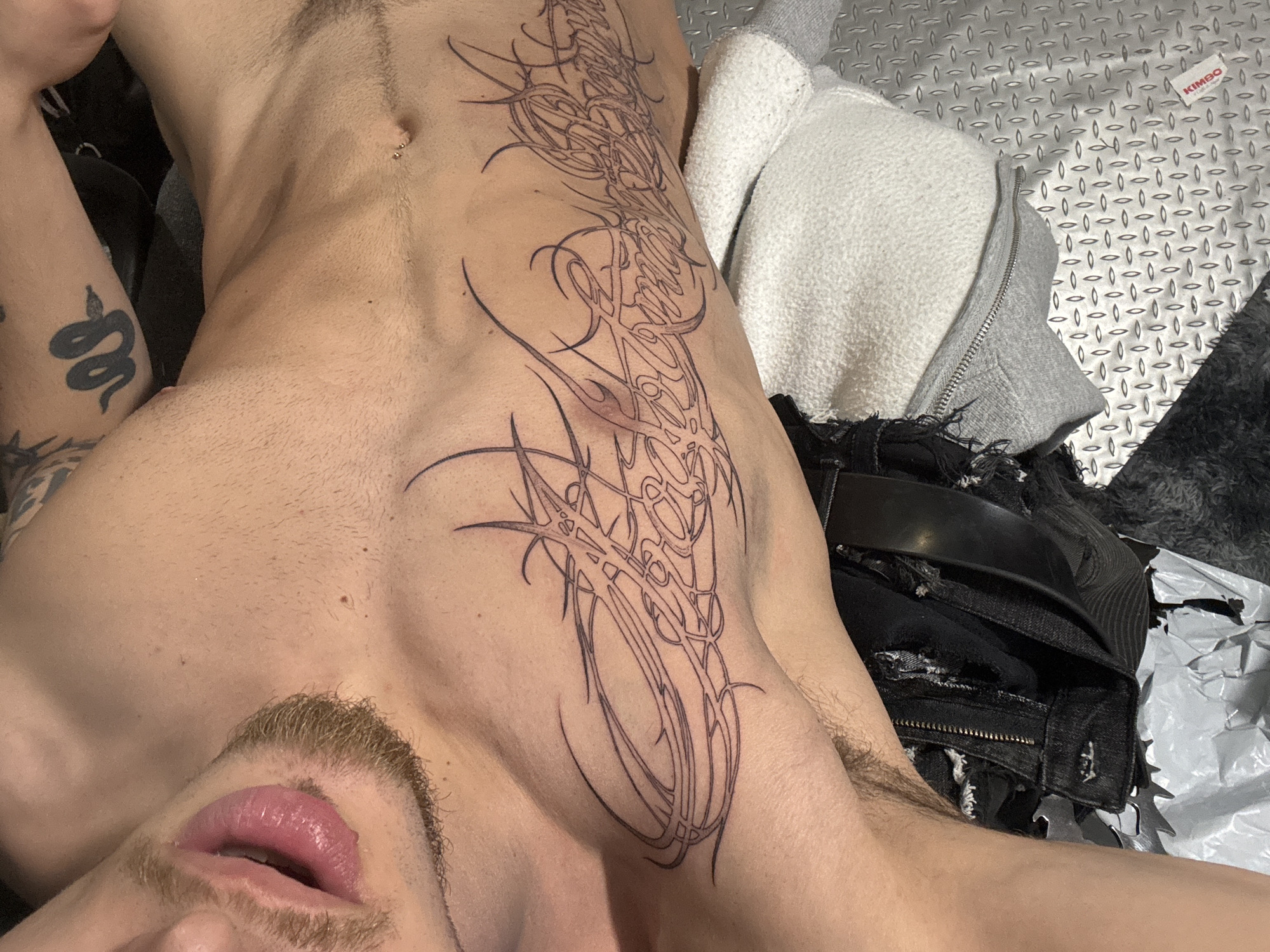 The image depicts a person lying on what appears to be a metallic surface with a patterned texture. The person has tattoos on their torso and arm. The torso tattoo is a large, abstract design extending from the chest to the abdomen. The arm tattoo depicts a snake. The person's face is partially visible, showing their mouth and mustache. A white and grey zipped hoodie is visible next to the person, as well as a pair of black jeans with a belt. The image doesn't provide any information about Sofia, Bulgaria.