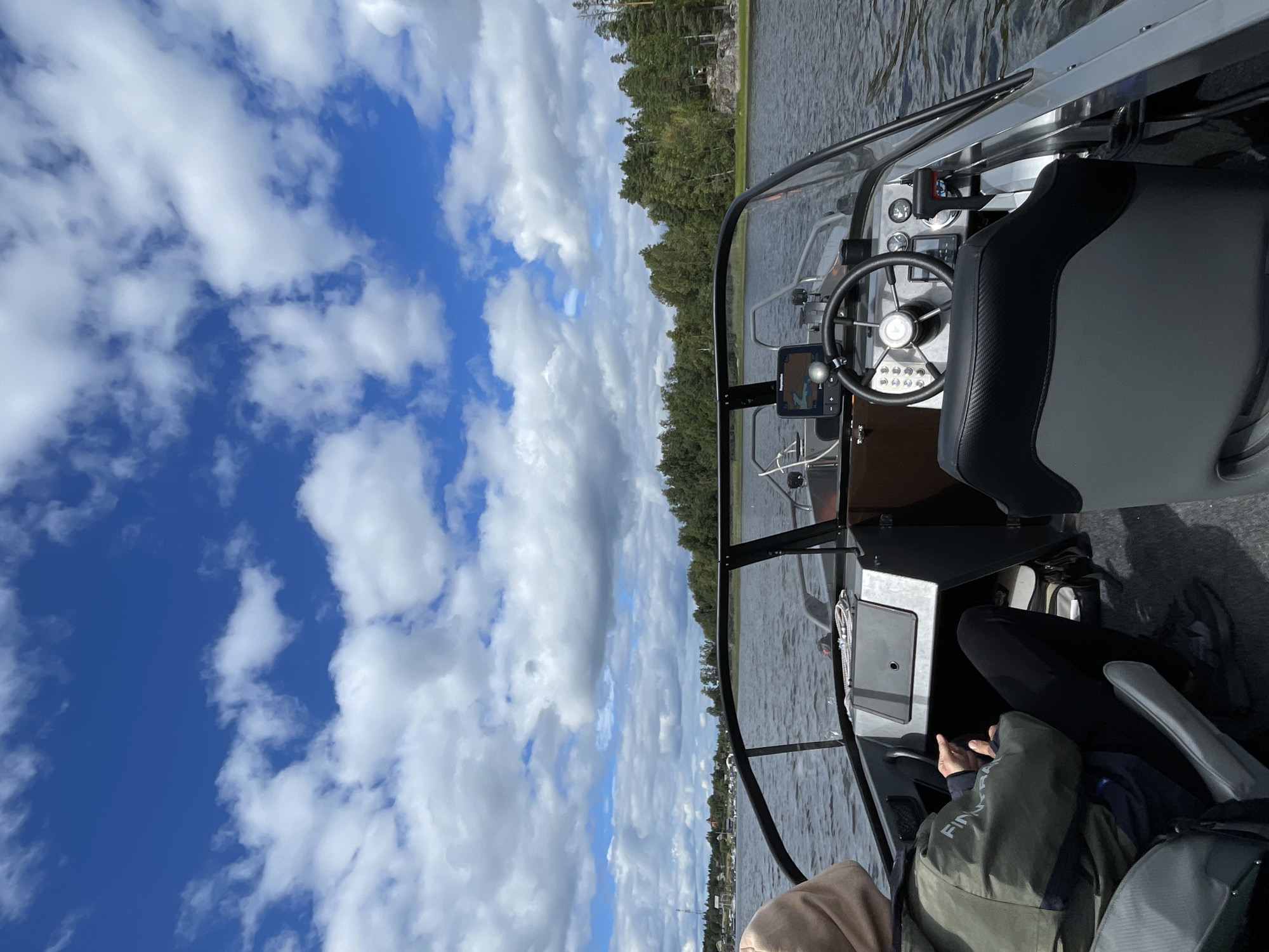 The image captures a view from inside a motorboat on a body of water under a cloudy blue sky. The boat's interior is visible, with a steering wheel, dashboard instruments, and seating. A person wearing a green jacket and a tan hood is seated in the boat, facing away from the camera. The background shows a tree-lined shore with some buildings visible in the distance. The scene appears to be a calm day on the water, possibly in a rural or suburban area. There is no indication of the city of Berozovo, Russia in this image.