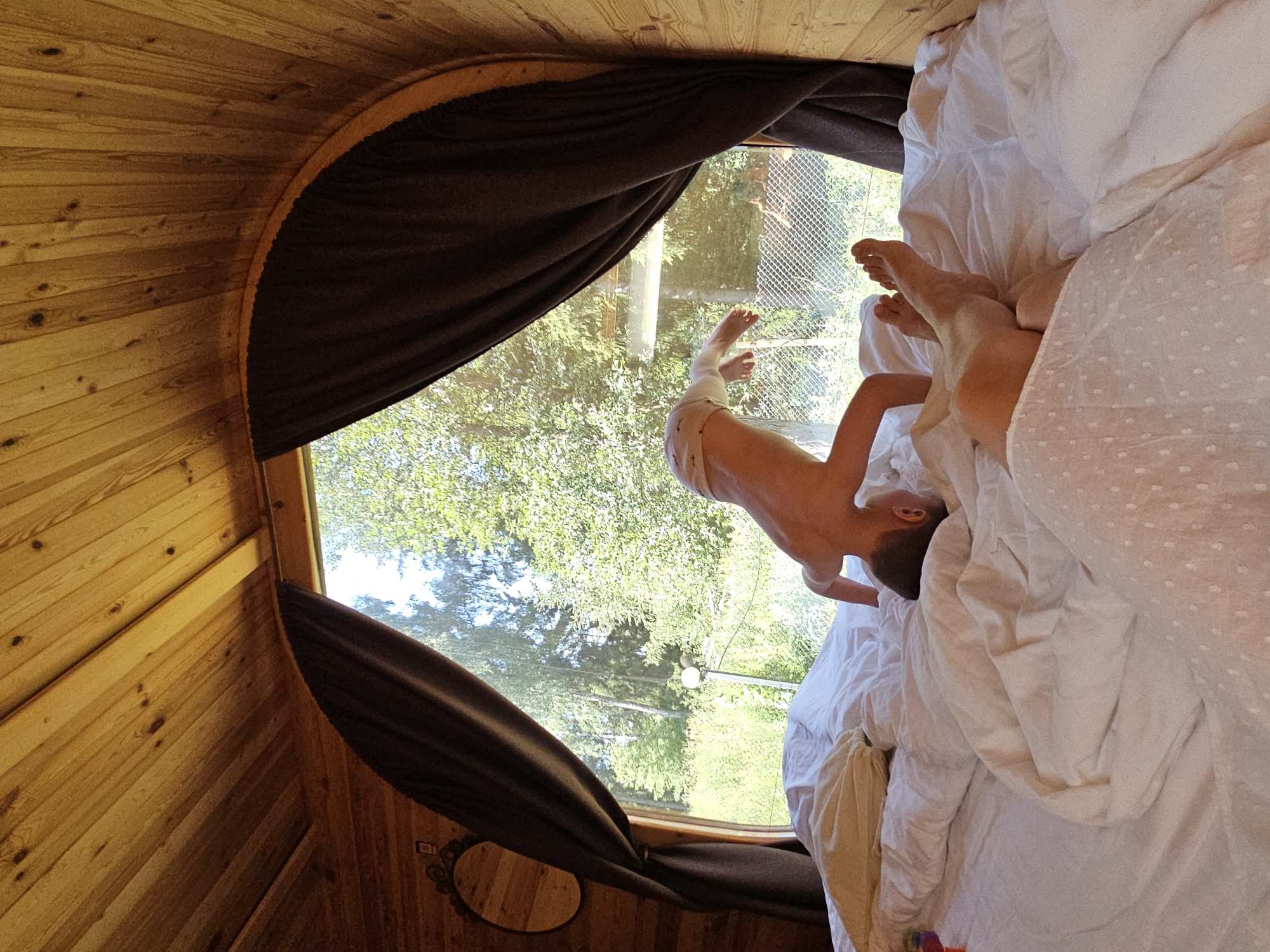 A boy is doing a handstand on a bed inside a wooden structure with a large arched window. Outside the window, green trees and vegetation are visible, along with a metal fence and a light post. The bed is covered in white bedding, and a person's legs are visible in the foreground. The wooden interior of the structure, likely a glamping pod, is also visible, with curved wooden planks forming the ceiling. The location is specified as Ulitkino, Russia.