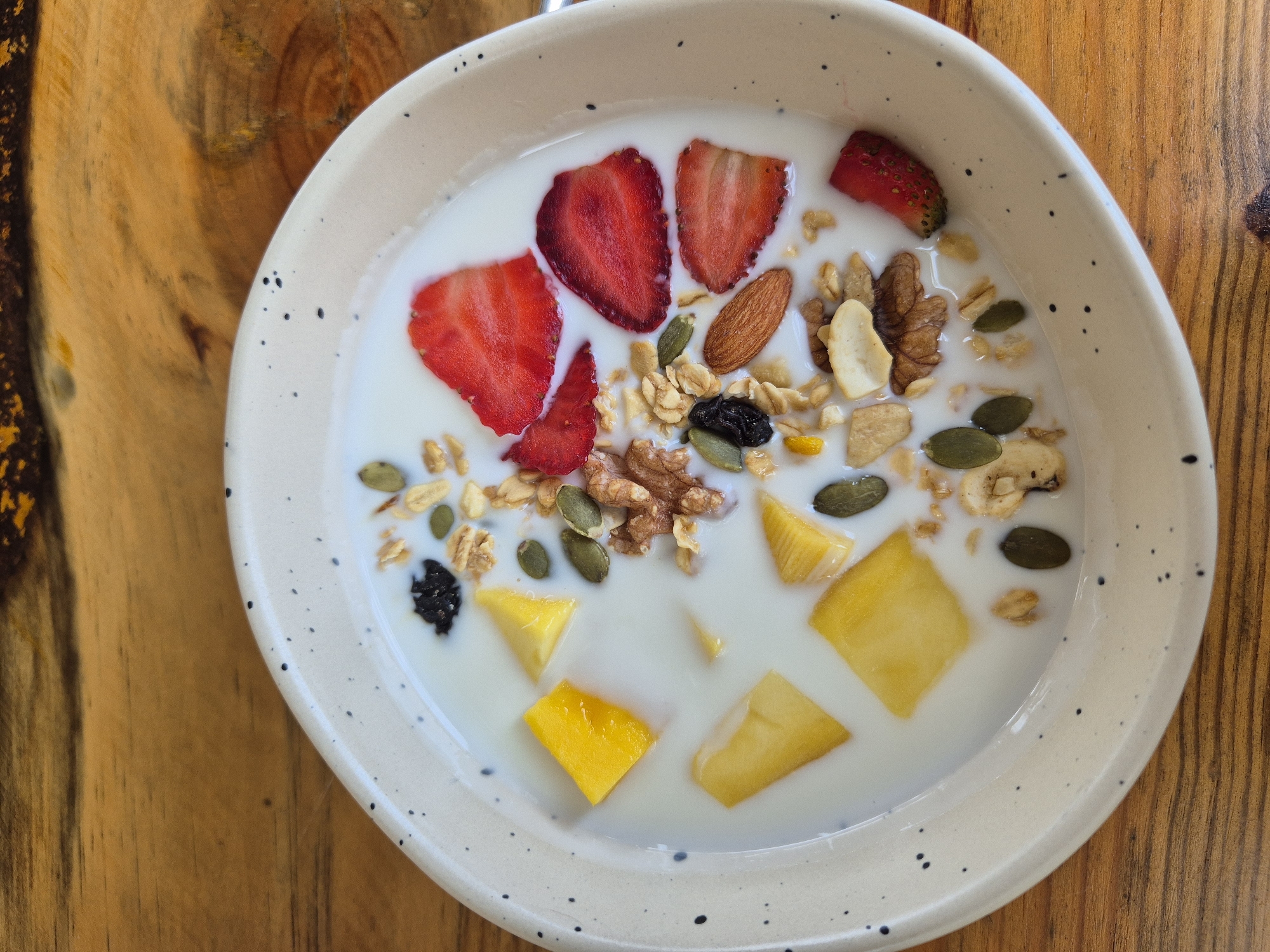 A close-up, top-down shot shows a bowl of yogurt with granola, strawberries, mango chunks, almonds, walnuts, pumpkin seeds, and raisins. The bowl is speckled white and sits on a wooden surface. There is no indication of a specific city or location within Vietnam.