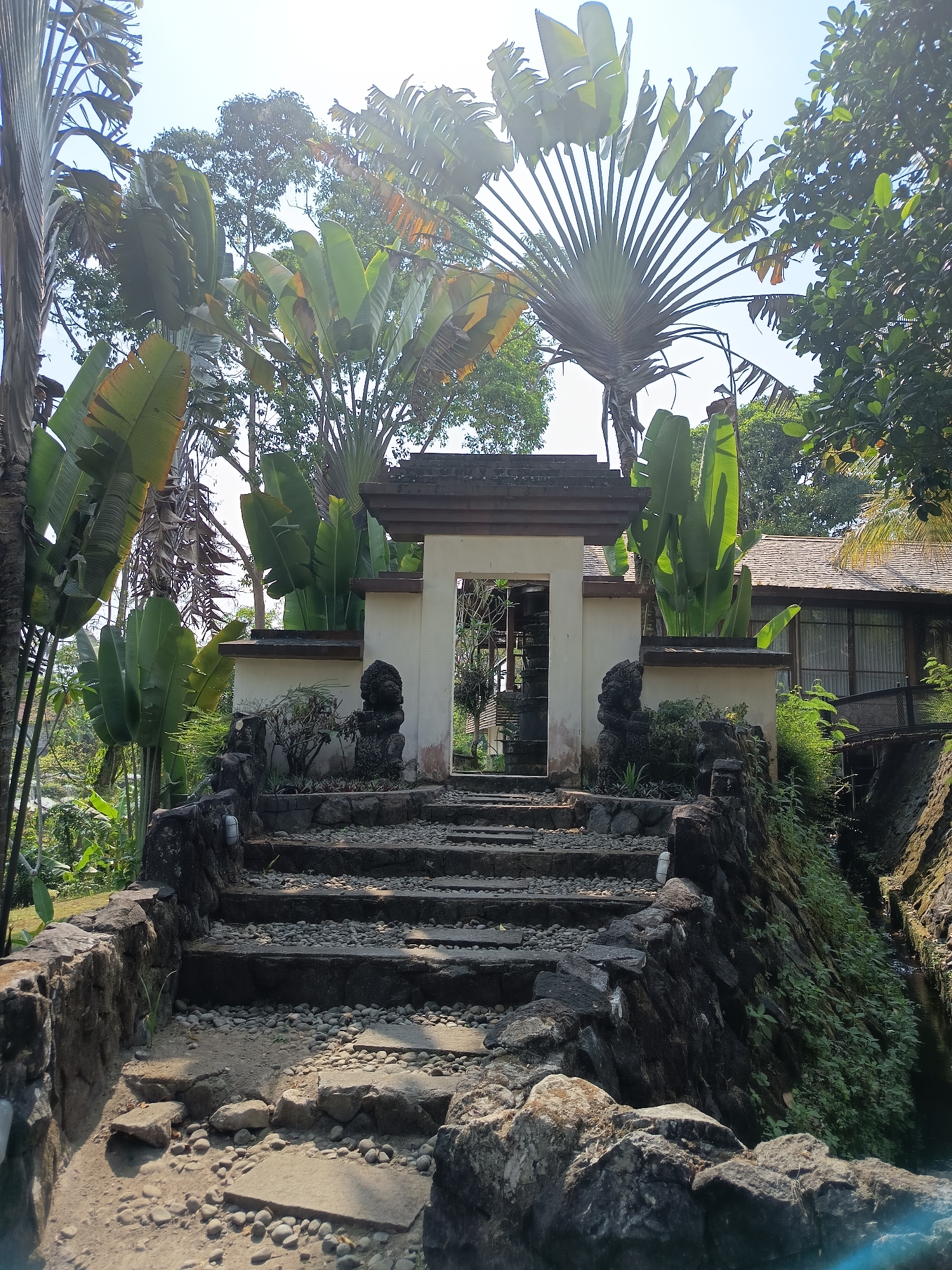 A stone staircase with pebble-filled treads and risers leads up to an arched entryway. The entryway is flanked by two dark stone lion statues. Beyond the entryway, lush green foliage, including large palm-like plants and trees, surrounds a building with a brown tiled roof. The scene is set outdoors, with a bright sky visible above the trees. The architectural style and dense tropical vegetation suggest the location is likely a resort or a similar establishment in a tropical climate, such as Ngaglik, Indonesia.