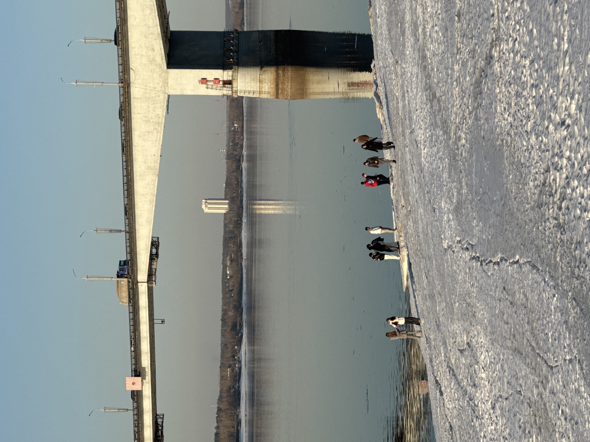 The image depicts a winter or early spring scene along the Kama River in Perm, Russia, under a clear, bright sky. The central feature is the Komunalny Bridge, a substantial concrete structure spanning the river. The bridge deck carries multiple traffic lanes, with vehicles, including a blue dump truck loaded with material and several cars, observed moving from left to right. Streetlights on tall poles are mounted along the bridge railings.
A massive concrete pier of the Komunalny Bridge dominates the right side of the frame, extending into the river and casting a long, dark shadow across the water and the snowy bank. Red warning markers are affixed to the pier.
On the foreground's snowy and icy bank, approximately ten individuals are present. They are dressed in winter or transitional outerwear such as coats and jackets.
- On the far left, two females stand near the water's edge, seemingly conversing. One wears a light-colored top and dark pants, facing another in a light brown coat.
- Moving right, a cluster of three individuals is observed, two in dark jackets and one in a white top, standing close together.
- Further right, another group of three includes a person in a red jacket walking right, accompanied by two others in dark jackets.
- On the far right, near the bridge pier, two individuals are standing: one in a brown jacket and another in a light jacket, observing the river.
The water of the Kama River is calm, reflecting the sky and distant structures. Patches of ice or slush are visible on the river's surface near the bank. The bank itself is covered in uneven, patchy snow and ice, with visible footprints.
The far bank of the river features a treeline, possibly dormant deciduous trees, with scattered buildings visible. A prominent, multi-sectioned white high-rise building, consistent with a landmark in Perm, stands on the distant horizon and is reflected in the river. Additional faint industrial or urban structures are visible further in the background. The sun is low, casting long shadows towards the left, indicating either morning or late afternoon.