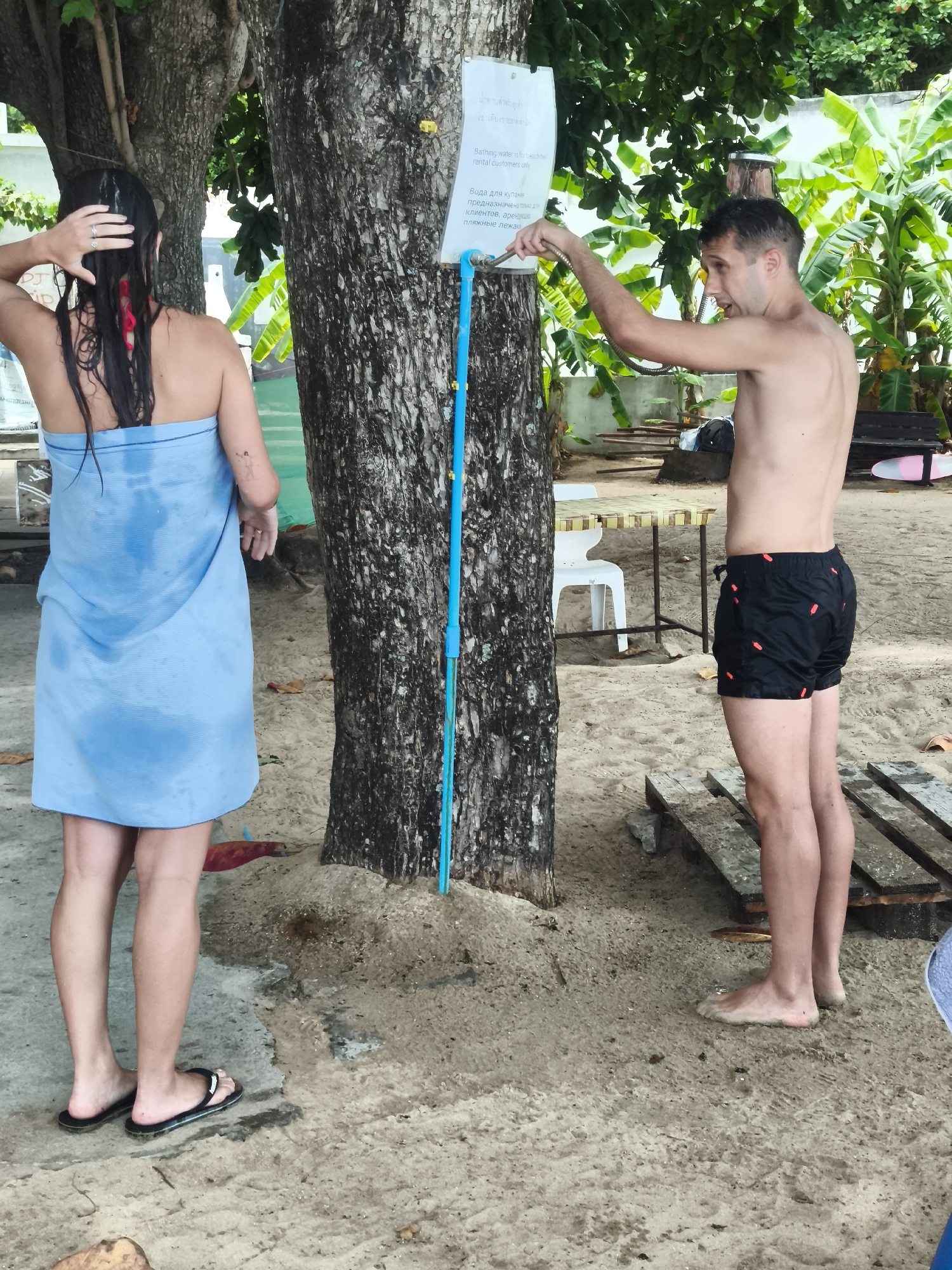 A man and a woman are standing at an outdoor shower on a sandy beach in Ko Samui District, Thailand. The woman, on the left, is facing away from the camera and is wrapped in a wet, light blue towel. She has long, dark hair and is wearing a red bikini top underneath the towel. She is wearing black flip-flops. The man, on the right, is facing the shower and is holding the showerhead, which is connected to a blue pipe attached to a tree. He is shirtless and wearing black swim trunks with a red pattern. The shower is positioned near the base of a large tree. In the background, there are more trees and foliage, as well as some tables and chairs, suggesting a beachfront establishment. The ground is sandy and wet in places, indicating recent use of the shower.