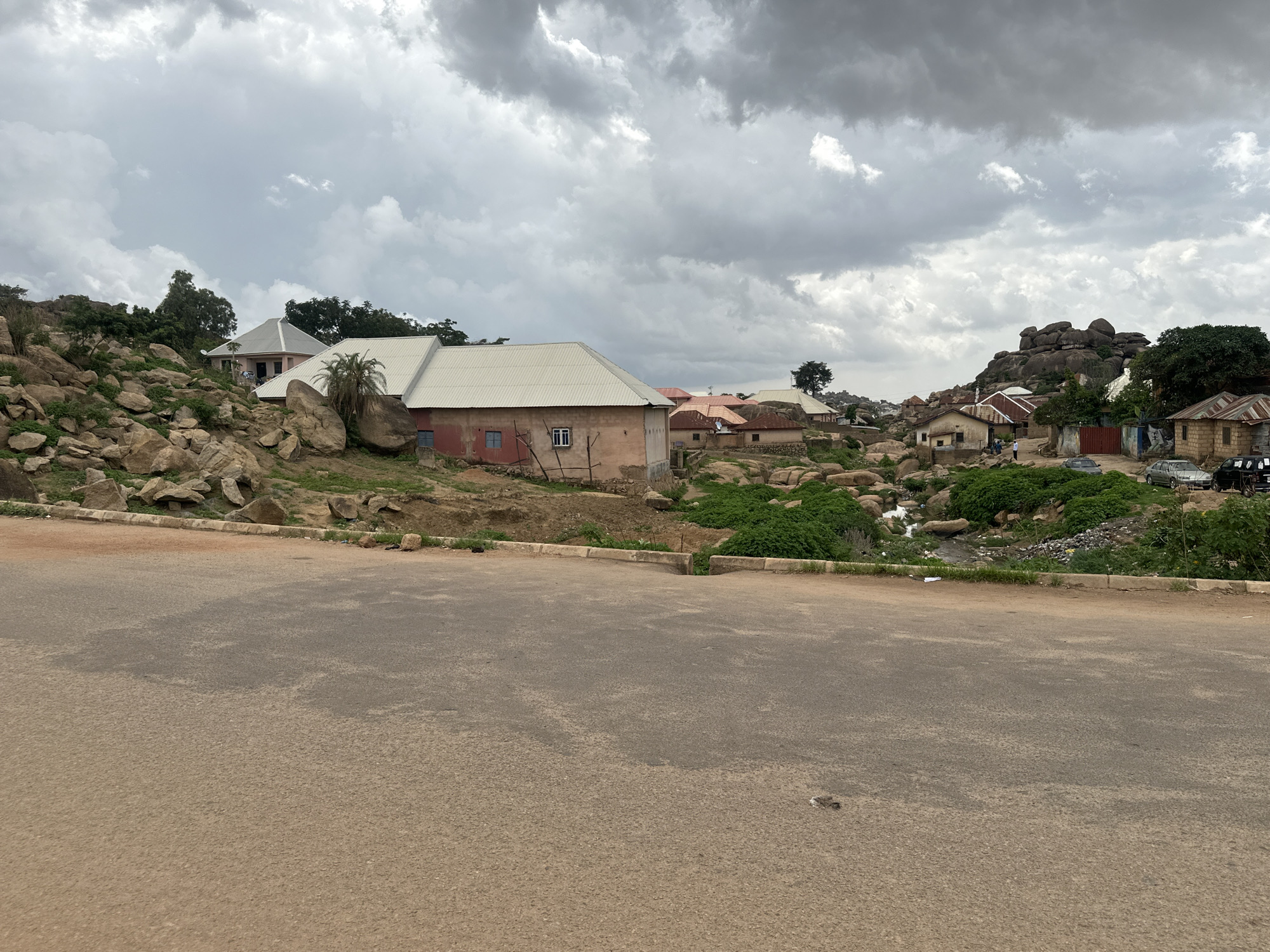 The image depicts a section of Jos, Nigeria, under a cloudy sky. A paved road dominates the foreground. Beyond the road, the terrain transitions to a mixture of rocks, vegetation, and residential buildings. The buildings vary in style, with some featuring corrugated iron roofs. A stream or drainage channel runs through the landscape, and trash is visible near it. Several parked cars can be observed in the distance, suggesting activity in the area.