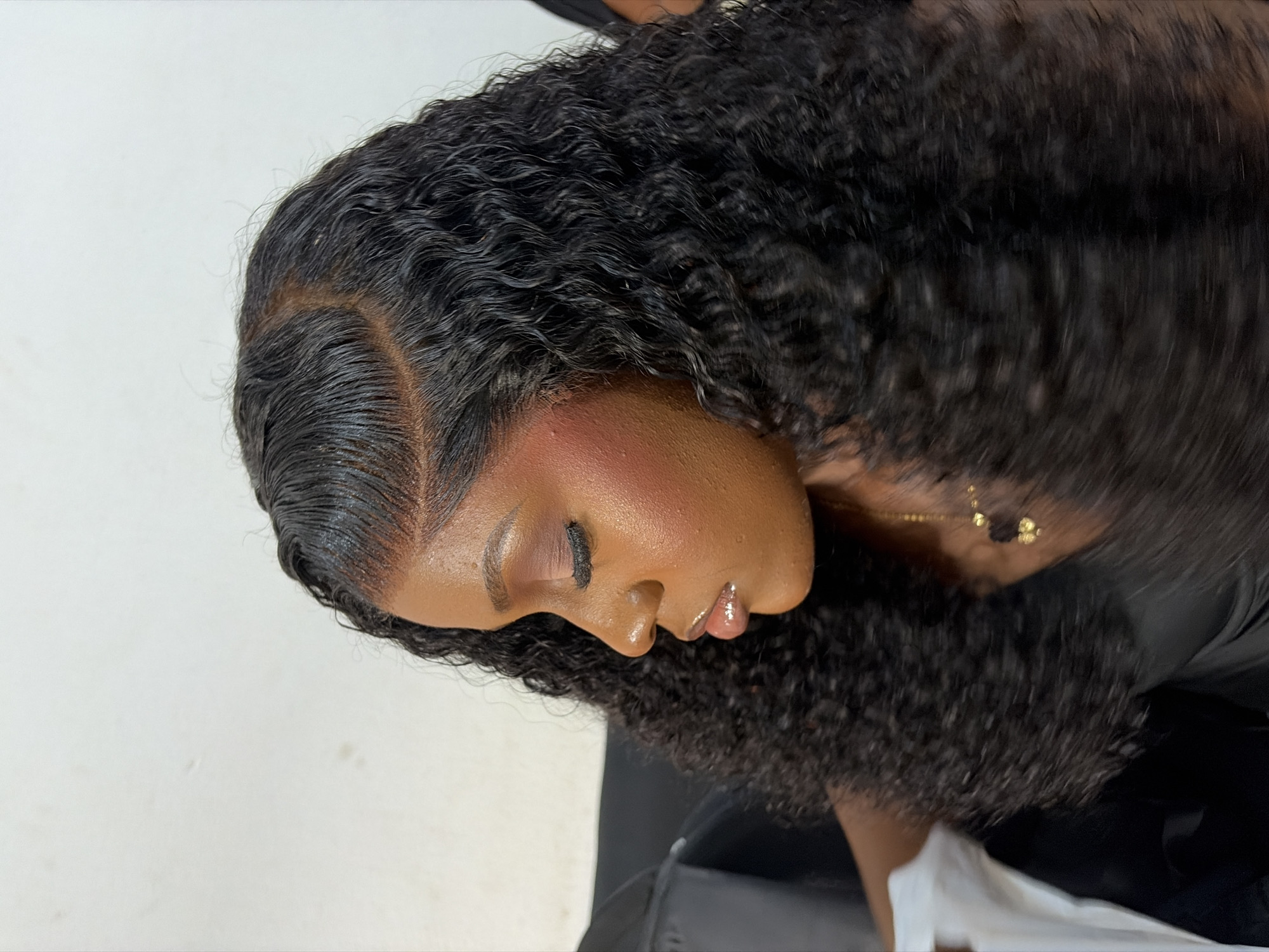 A single individual, a young adult female with dark skin, is depicted in a close-up, right-profile photographic portrait, with eyes closed or cast downwards. Her dark, curly hair, styled with a defined side part and laid baby hairs along the hairline, exhibits a wet appearance. She wears full facial makeup, including reddish-brown eyeshadow, prominent dark false eyelashes, reddish-orange blush on her cheeks, and glossy, light-colored lips. A delicate gold-colored chain with two small, dark, possibly heart-shaped pendants is visible around her neck. She appears to be wearing a dark garment. The background consists of a plain, light-colored wall, with a darker, indistinct object partially visible on the far left. The scene is located in Gura Top, Nigeria.
