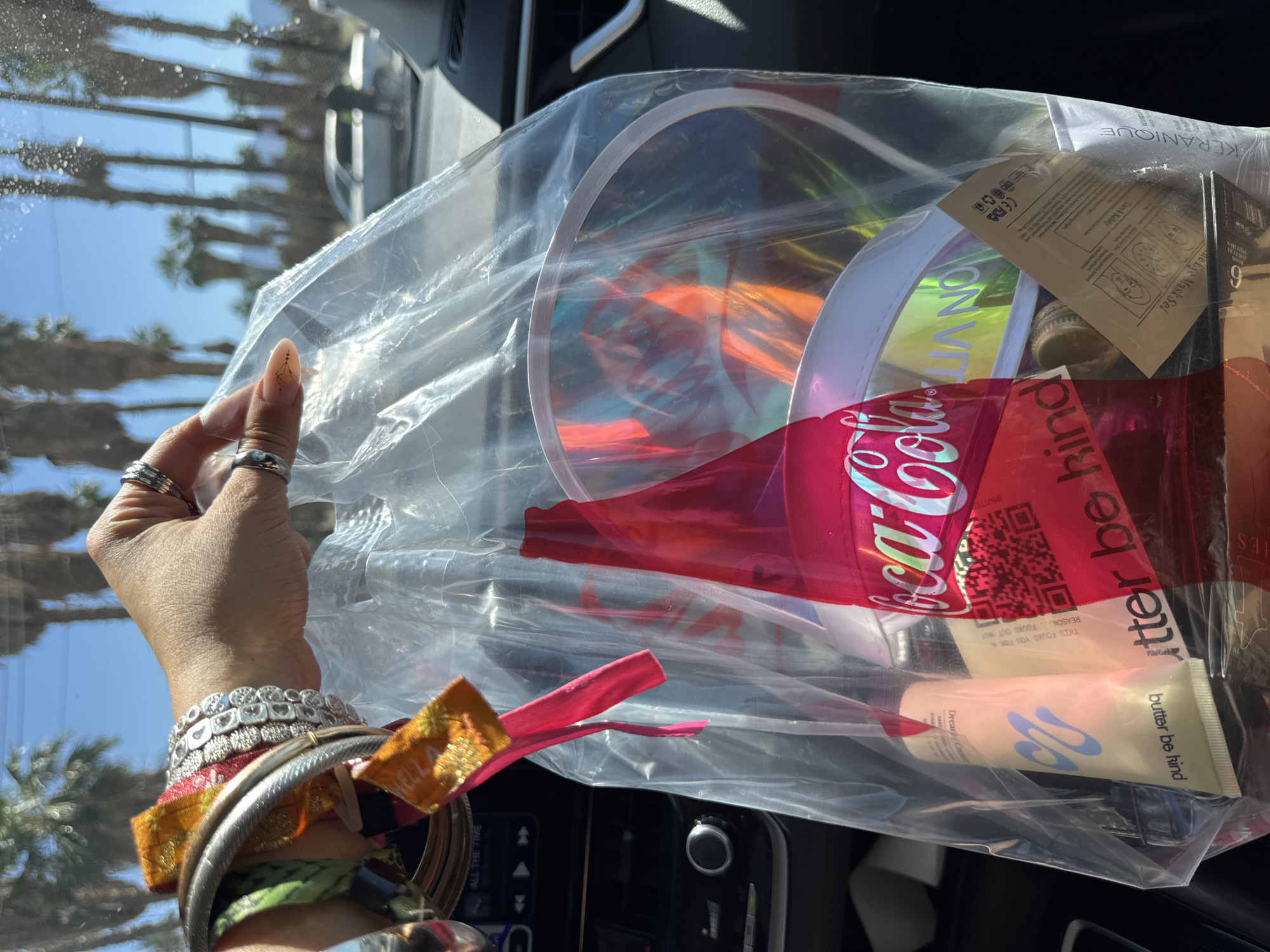A person inside a vehicle is holding up a plastic bag containing various items. The contents include a Coca-Cola branded item, a tube of "butter be kind" lotion, and a Keranique hair product. The person is wearing multiple bracelets and rings. Through the car window, palm trees lining a street are visible, suggesting the scene is in Thermal, United States of America.