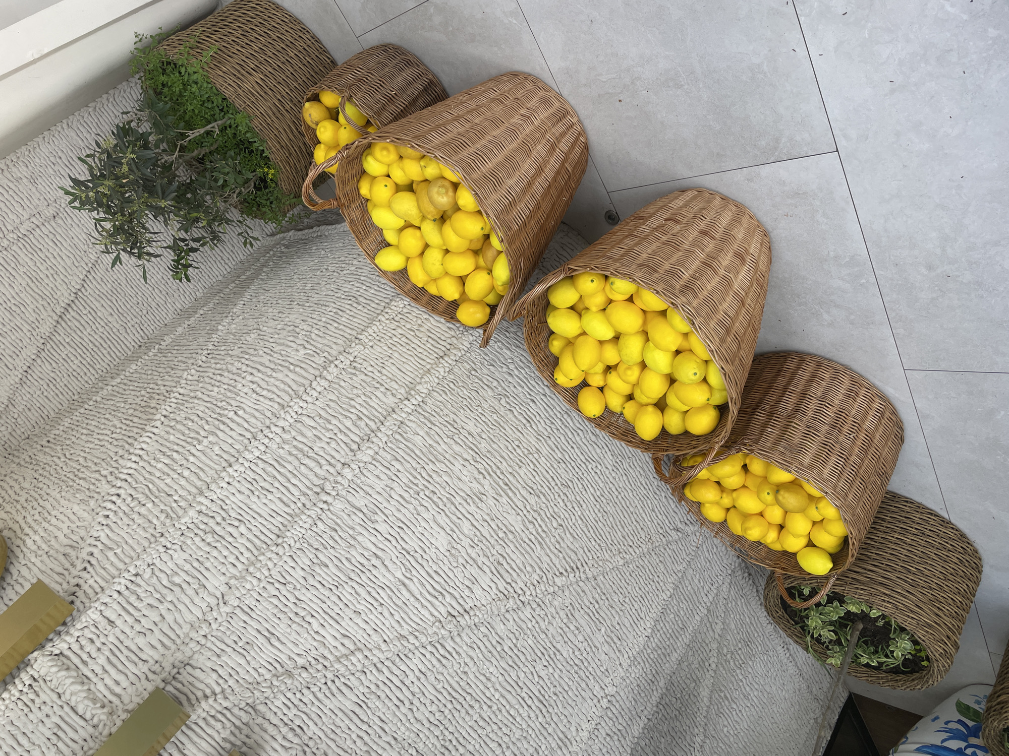 The image shows an indoor setting with a light gray tiled floor and a white textured rug. Several woven baskets are scattered across the rug, some filled with lemons and others with greenery. The environment appears to be a display or decorative arrangement, possibly in a store or studio. There are no people visible. The image does not provide enough information to confirm the location in Sochi, Russia.