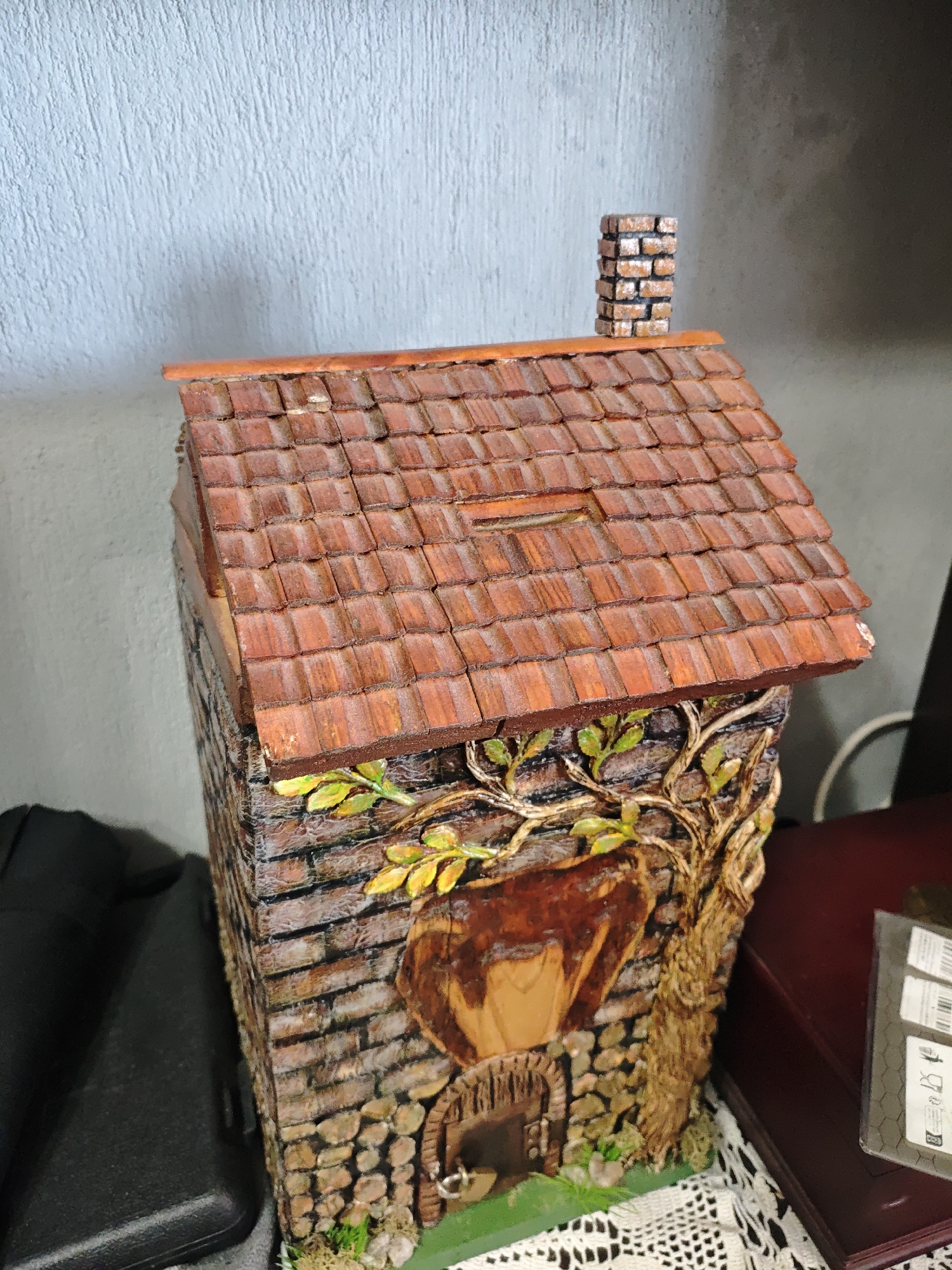 A miniature diorama of a house is set indoors. The house has a brick facade, a red tile roof with a small chimney, and a tree on one side. The house sits on a white crocheted surface and is positioned near a red-brown wooden box. To the left of the house is a black rectangular object. The background consists of a light blue wall. The city of Unknown and Unknown are not known to be where this image was taken.