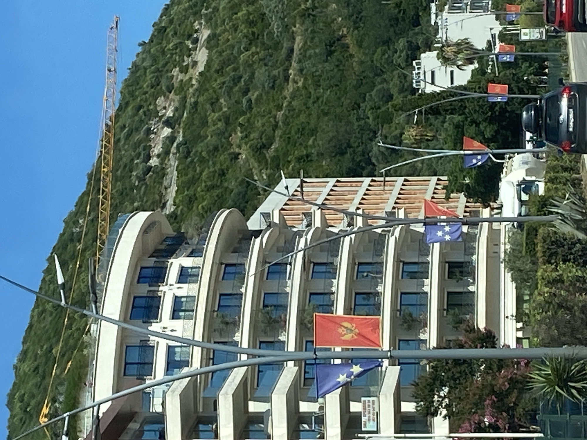 This image shows a street scene in Budva, Montenegro, with a focus on a modern, multi-story building featuring curved balconies and large windows. Several flags are displayed on poles in front of the building; prominently featured are the flag of Montenegro and a blue flag with stars, likely representing the European Union. A construction crane is visible against a green, forested hillside in the background, suggesting ongoing development in the area. Vehicles, including a dark-colored car with a rooftop carrier, are present on the road. The environment is sunny with a clear blue sky.
