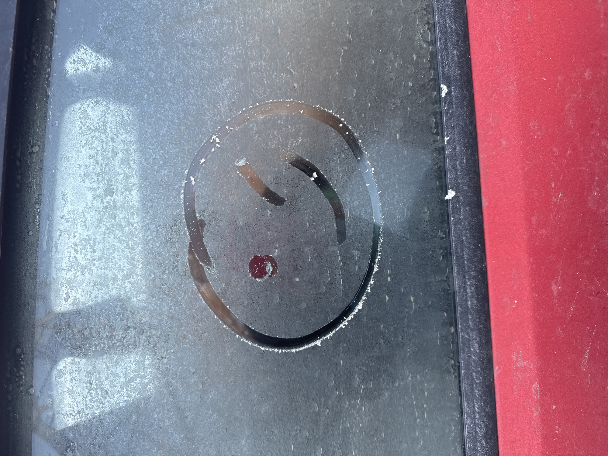 The image depicts a crudely drawn smiley face on a frosted window, likely of a vehicle. The window is bordered by a dark, possibly black, rubber or plastic frame. To the right of the window frame is a red surface, possibly the body panel of a car. The frost on the window appears uneven, with some areas wiped clear to form the smiley face. The smiley face consists of a circle, two curved lines representing eyes, and a circular mark, which could be intended to represent a nose. The overall scene appears to be outdoors, given the frost formation. There is no information in the image to determine if it is located in the city of Chaikovsky, Russia.
