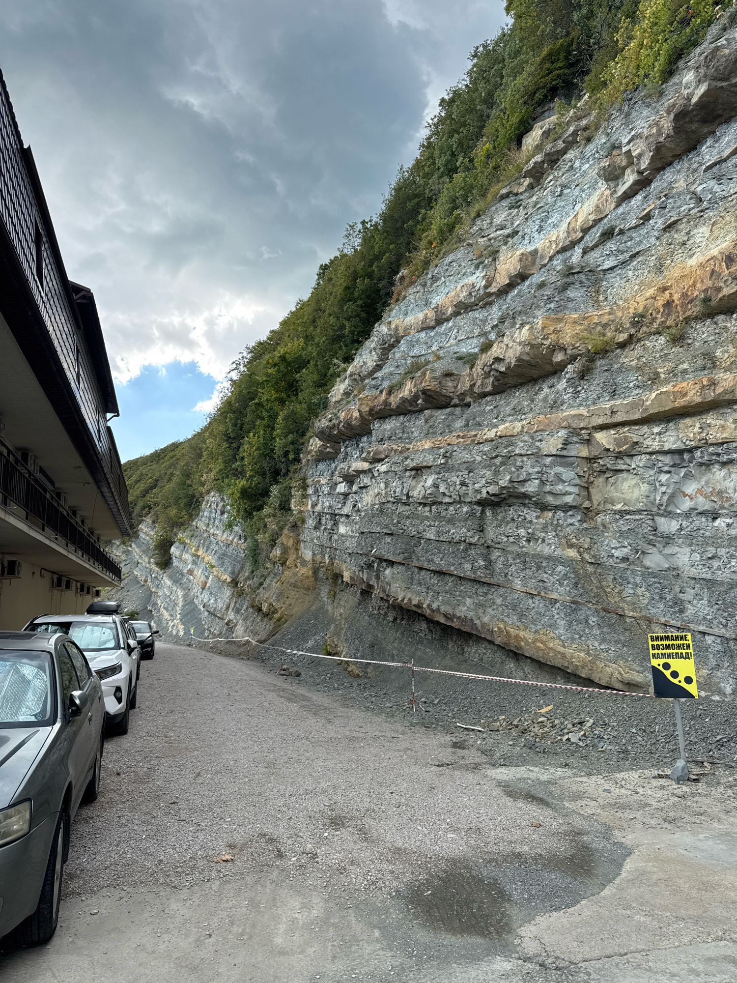 A gravel road runs alongside a sheer rock face in Novomikhaylovsky, Russia. The rock face exhibits distinct horizontal striations and is covered in vegetation. A multi-story building with balconies and air conditioning units lines the left side of the road. Several cars are parked along the right side of the road. A yellow warning sign with black circles depicting falling rocks is posted near the rock face, along with a red and white striped barrier tape. The sky is overcast.